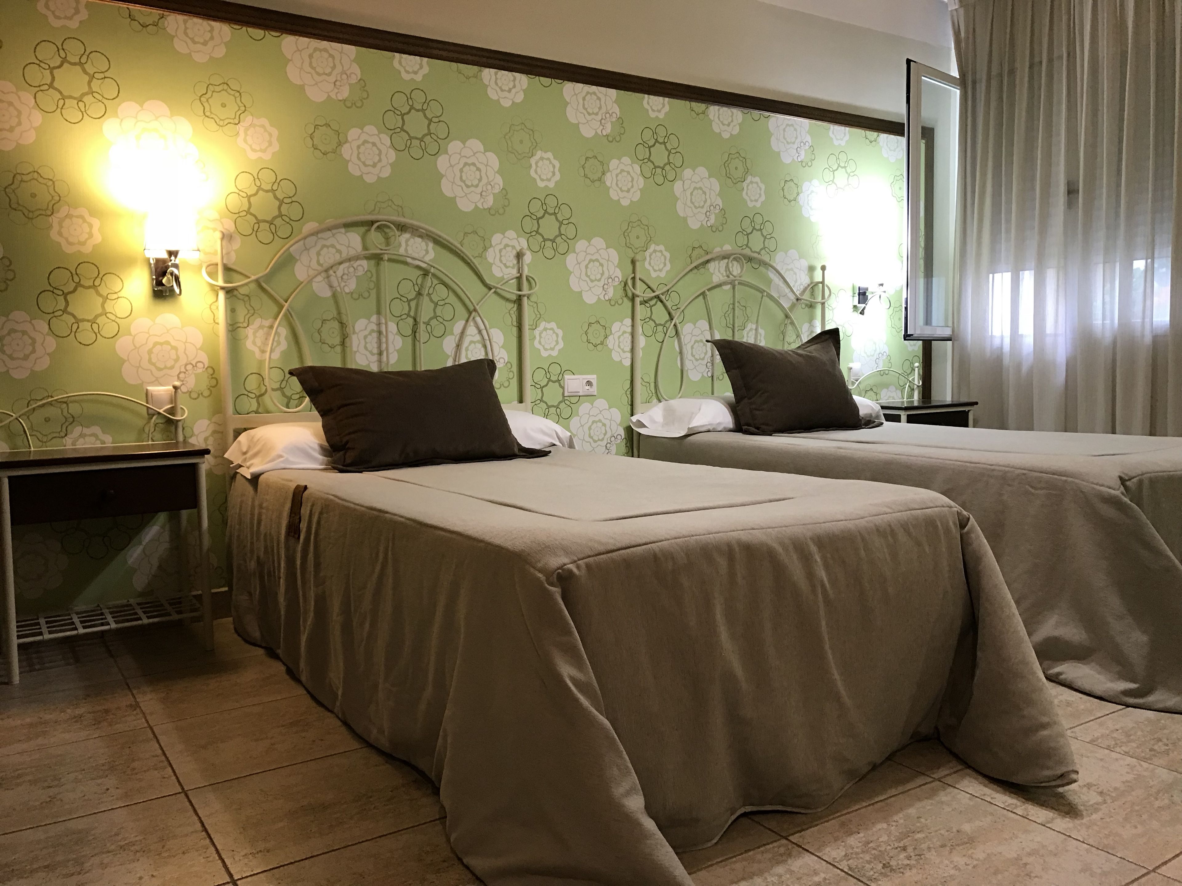 A hotel room with two beds, green floral wallpaper, a nightstand with a lamp, and a TV on the wall.