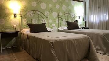 A hotel room with two beds, green floral wallpaper, a nightstand with a lamp, and a TV on the wall.