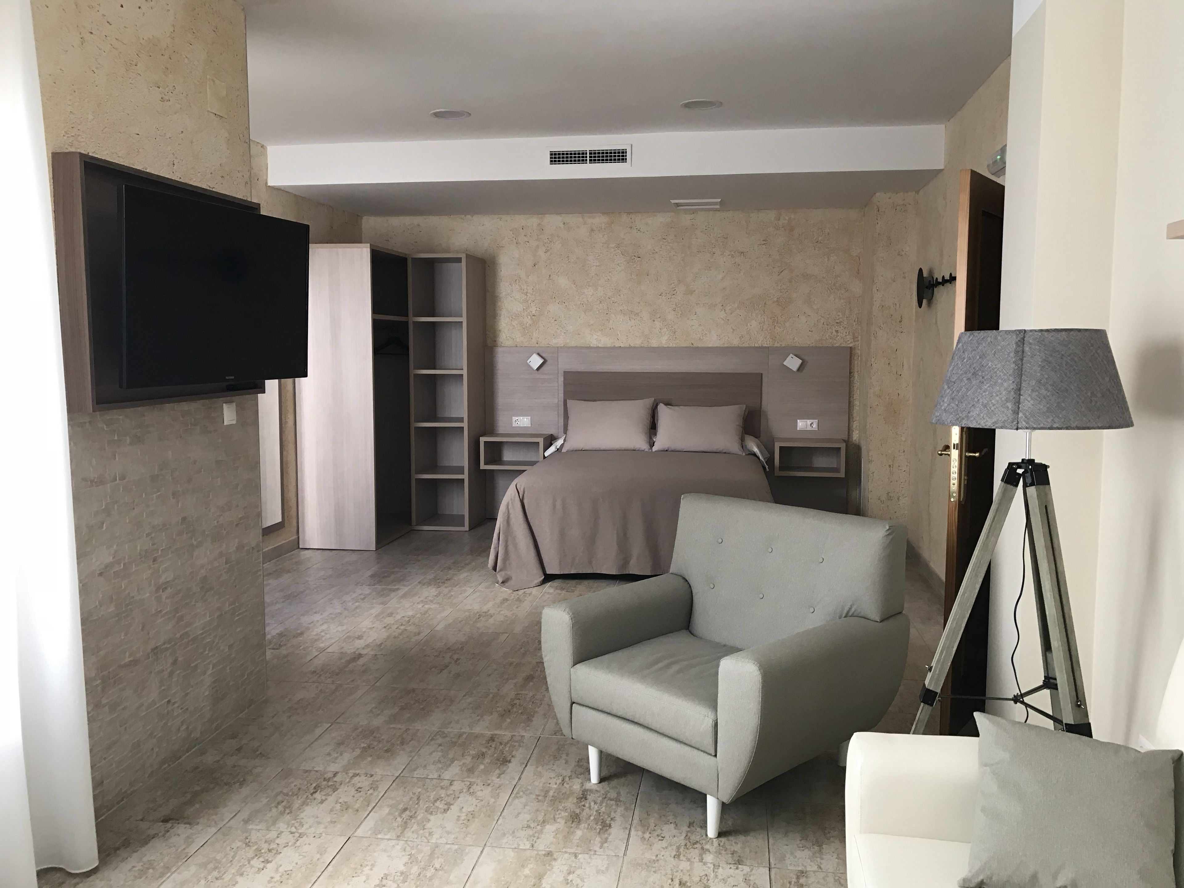 A modern hotel room with a bed, a grey armchair, a bedside table, a lamp, and a TV mounted on the wall.