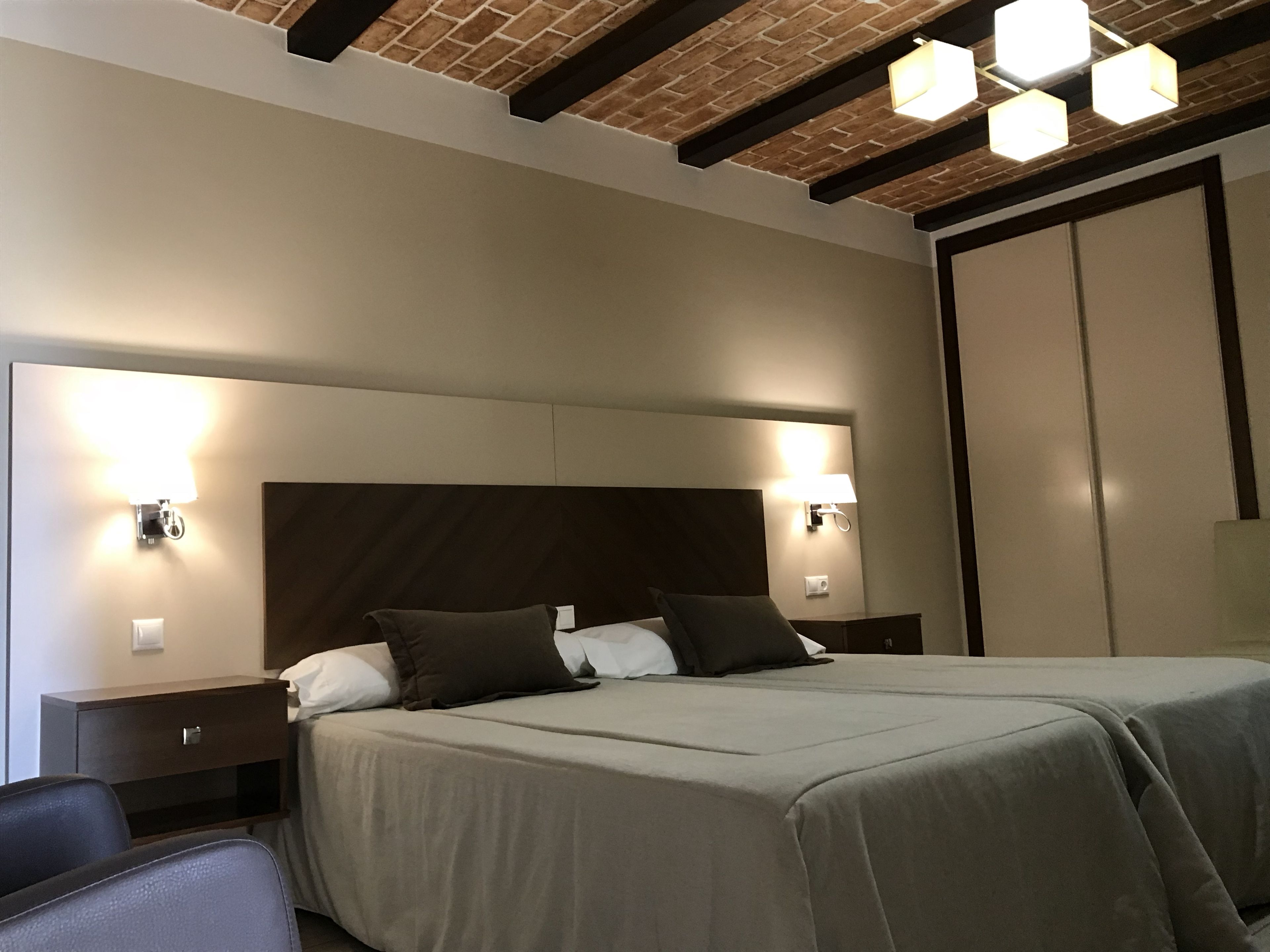 Habitación de hotel con una cama grande, dos mesitas de noche, un armario y un techo con ladrillo visto e iluminación empotrada.