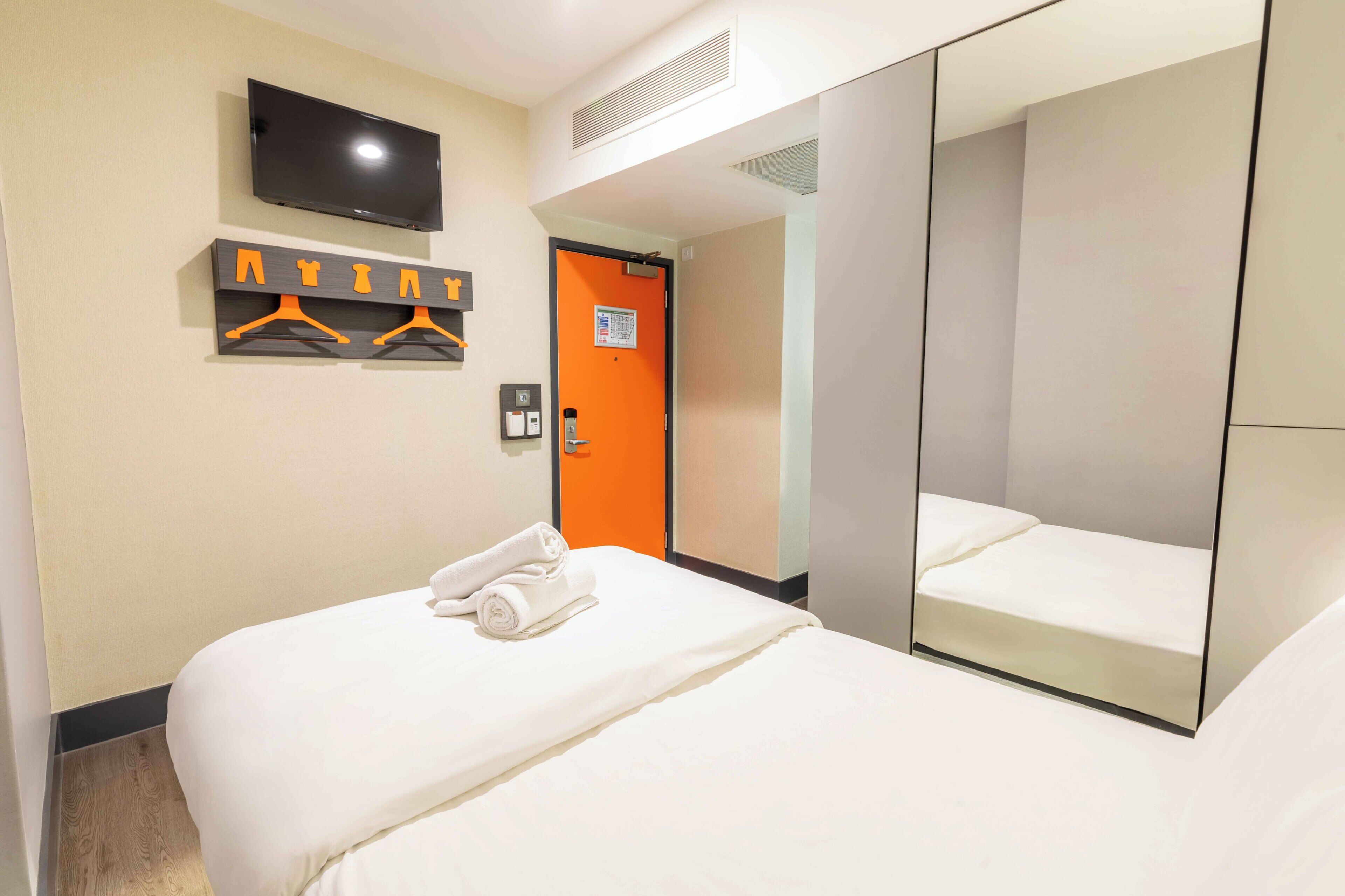 Habitación de hotel con cama, televisor montado en la pared, espejo y puerta con marco naranja.