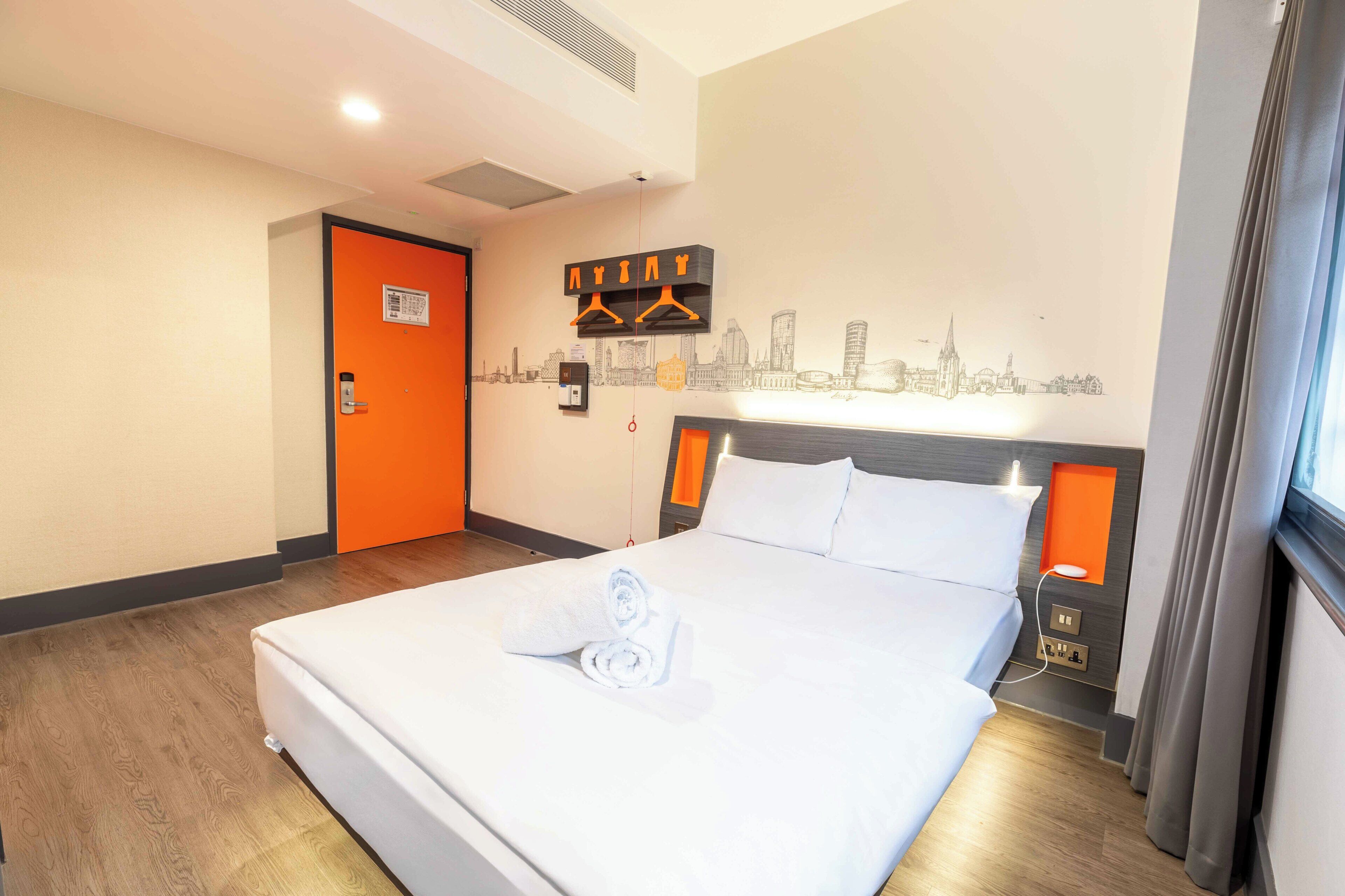 A hotel room with a bed, a cityscape wall art, a door, and a window with curtains.
