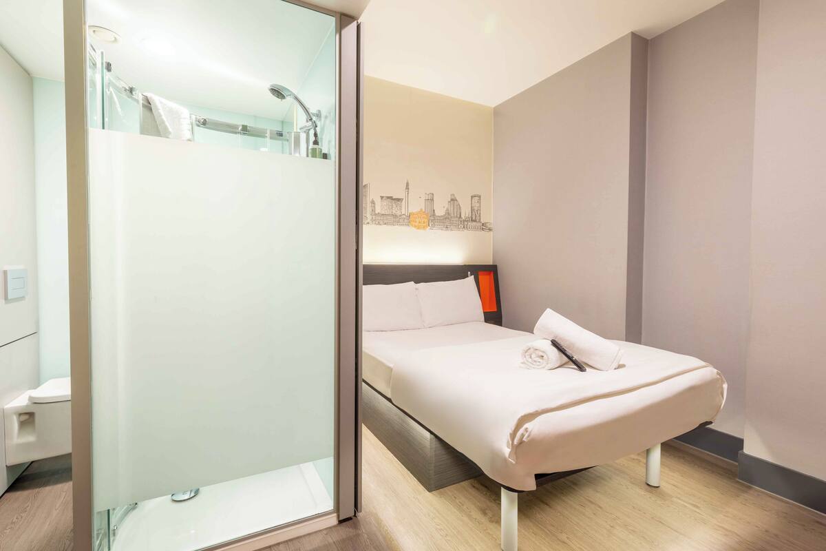 Ein modernes Hotelzimmer mit Duschkabine aus Glas, einem Bett mit Stadtbild-Wandgemälde und einem Nachttisch mit Lampe.