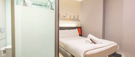 A modern hotel room with a glass shower enclosure, a bed with a cityscape mural on the wall, and a bedside table with a lamp.