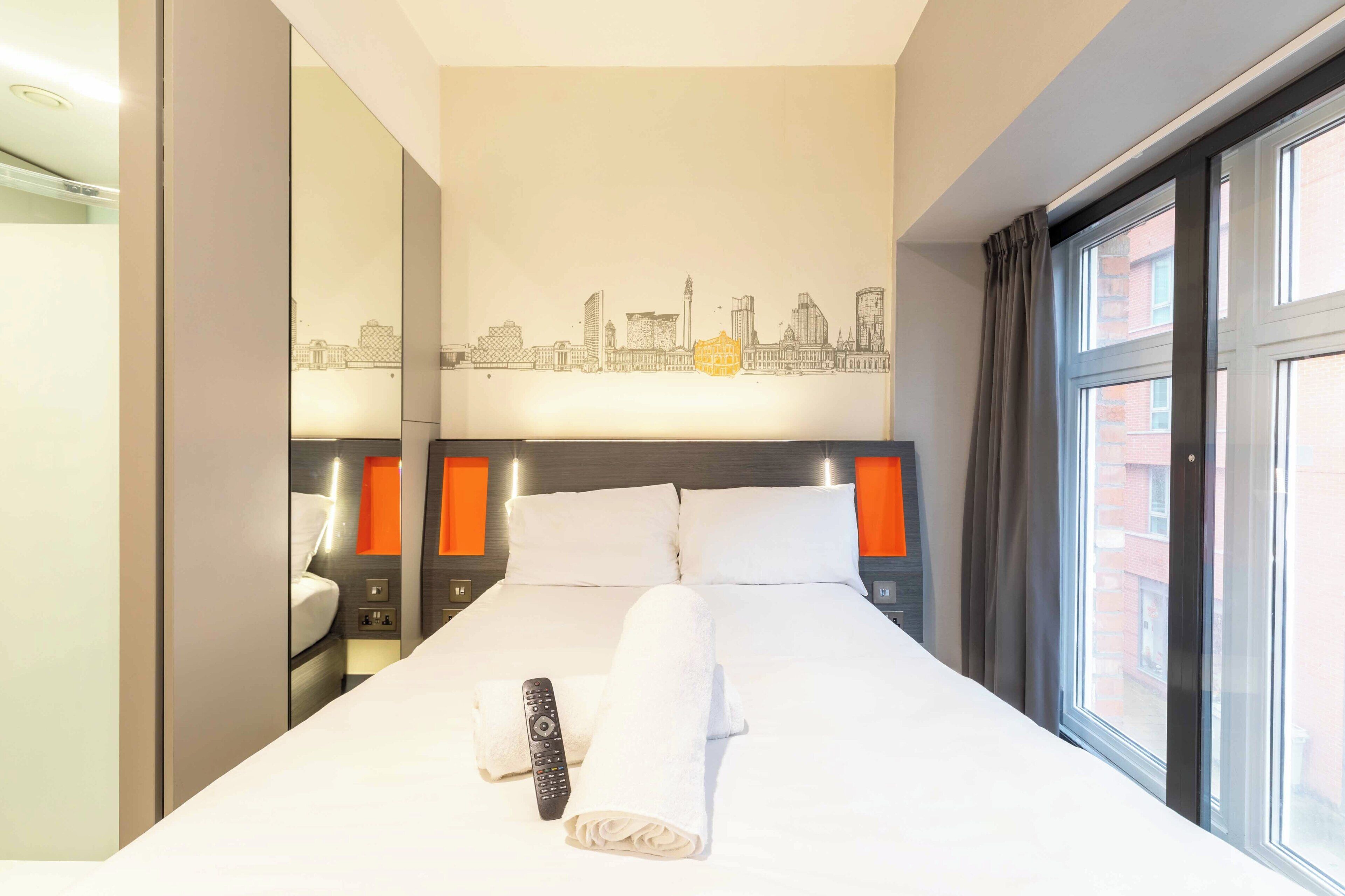 A hotel room with a bed, a cityscape mural, a window with curtains, and a nightstand with a remote control.