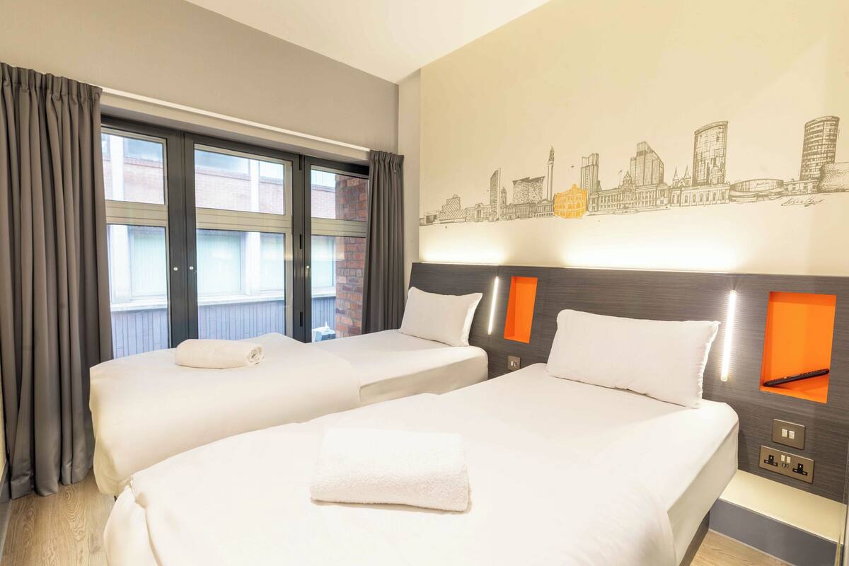 Ein Hotelzimmer mit zwei Betten, einer Stadtlandschaft als Wandbild und einem Fenster mit Vorhängen.