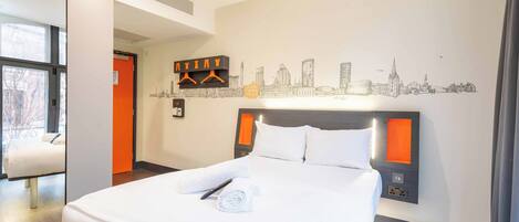 A hotel room with a large bed, a cityscape wall art, a coat rack, and a window with a view.