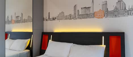 Una camera d'albergo con un letto grande, una testiera illuminata e un murale con una veduta urbana, oltre a uno specchio che riflette l'ambiente.