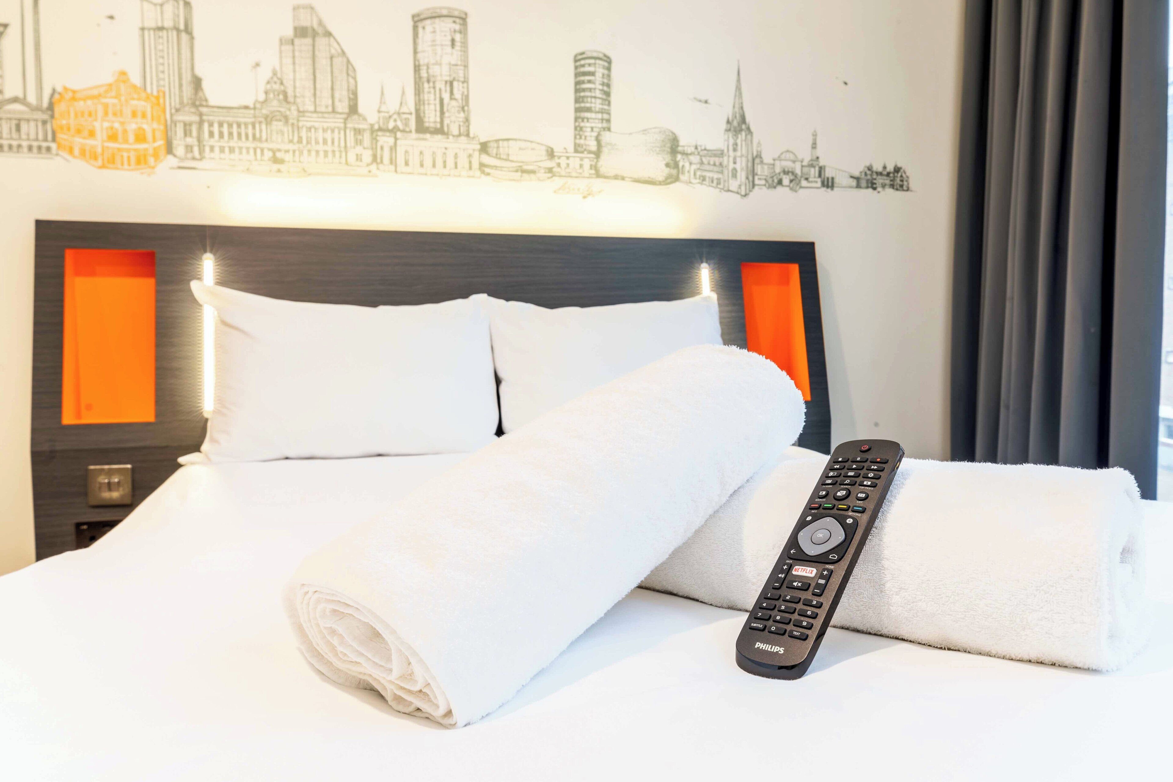Ein Hotelzimmer mit einem ordentlich bezogenen Bett, einer Fernbedienung und einem Stadtbild an der Wand.