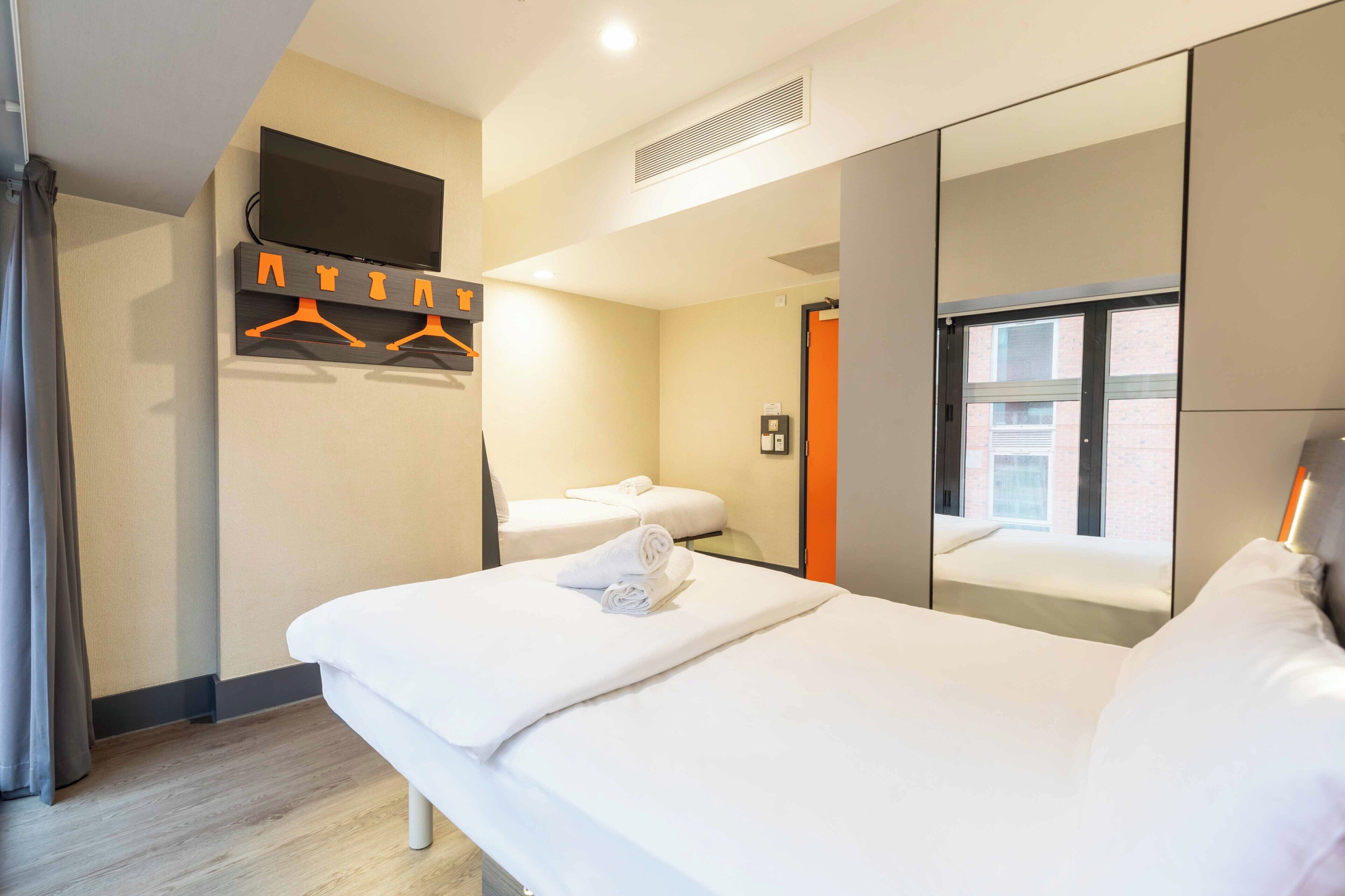 A hotel room with a bed, a TV mounted on the wall, a mirror, and a window.