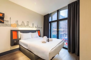 A hotel room with a bed, a cityscape wall art, a window with curtains, and a view of buildings outside. - easyHotel Birmingham (Birmingham)