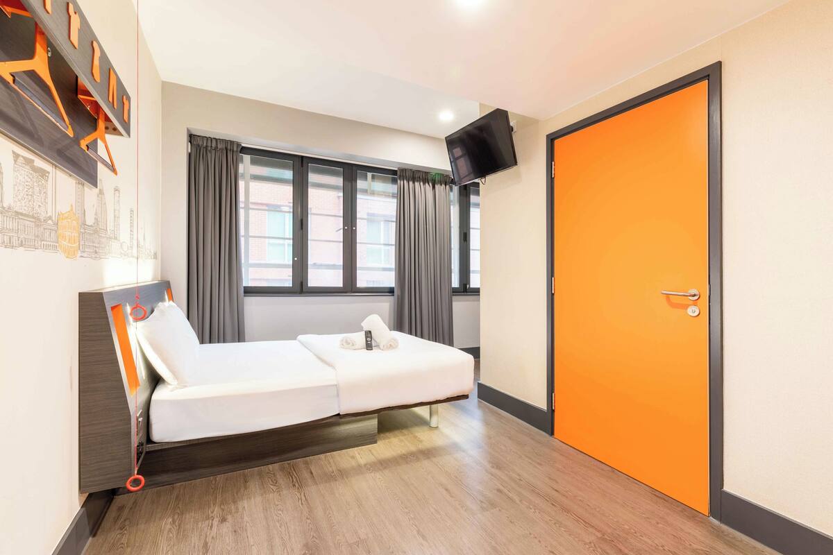 Ein Zimmer mit einem Einzelbett, Holzboden, einem großen Fenster mit Vorhängen, einem an der Wand montierten Flachbildfernseher und einer orangen Tür.