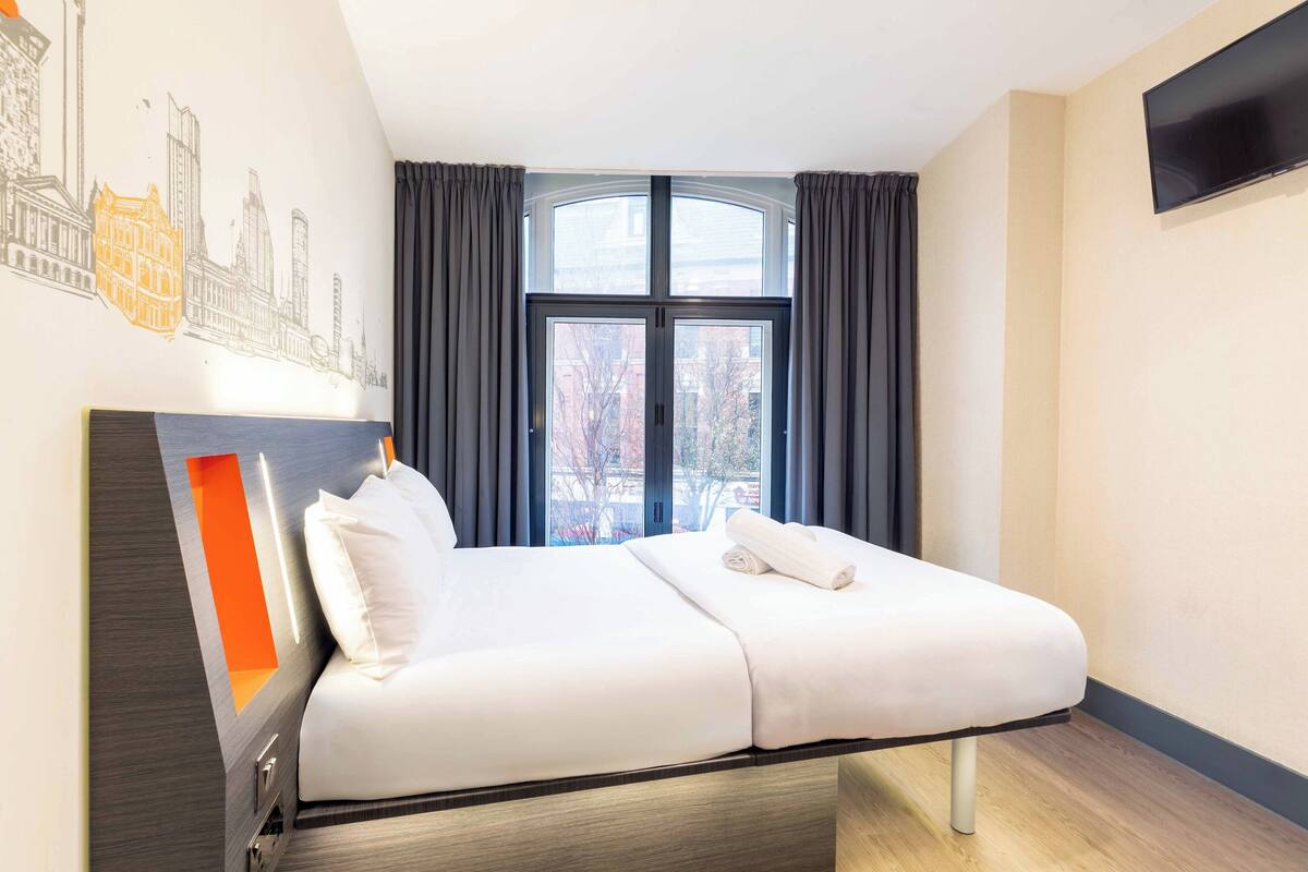 Ein Hotelzimmer mit Bett, einem Fenster mit Vorhängen, einem an der Wand befestigten Fernseher und einem Stadtbild-Wandgemälde.