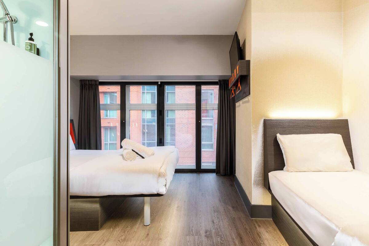 Ein Hotelzimmer mit zwei Betten, einem großen Fenster mit Vorhängen, einem Holzboden und einem an der Wand befestigten Flachbildfernseher.