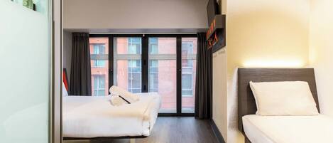 A hotel room with two beds, a large window with curtains, a wooden floor, and a flat-screen TV mounted on the wall.