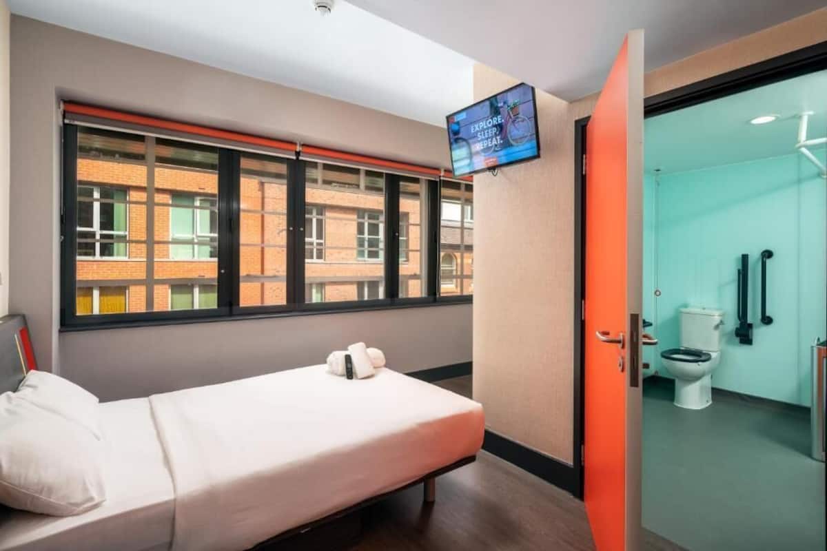 Ein Hotelzimmer mit Bett, Wandfernseher, Fenster mit Blick auf Gebäude und einem Badezimmer mit Toilette und Dusche.