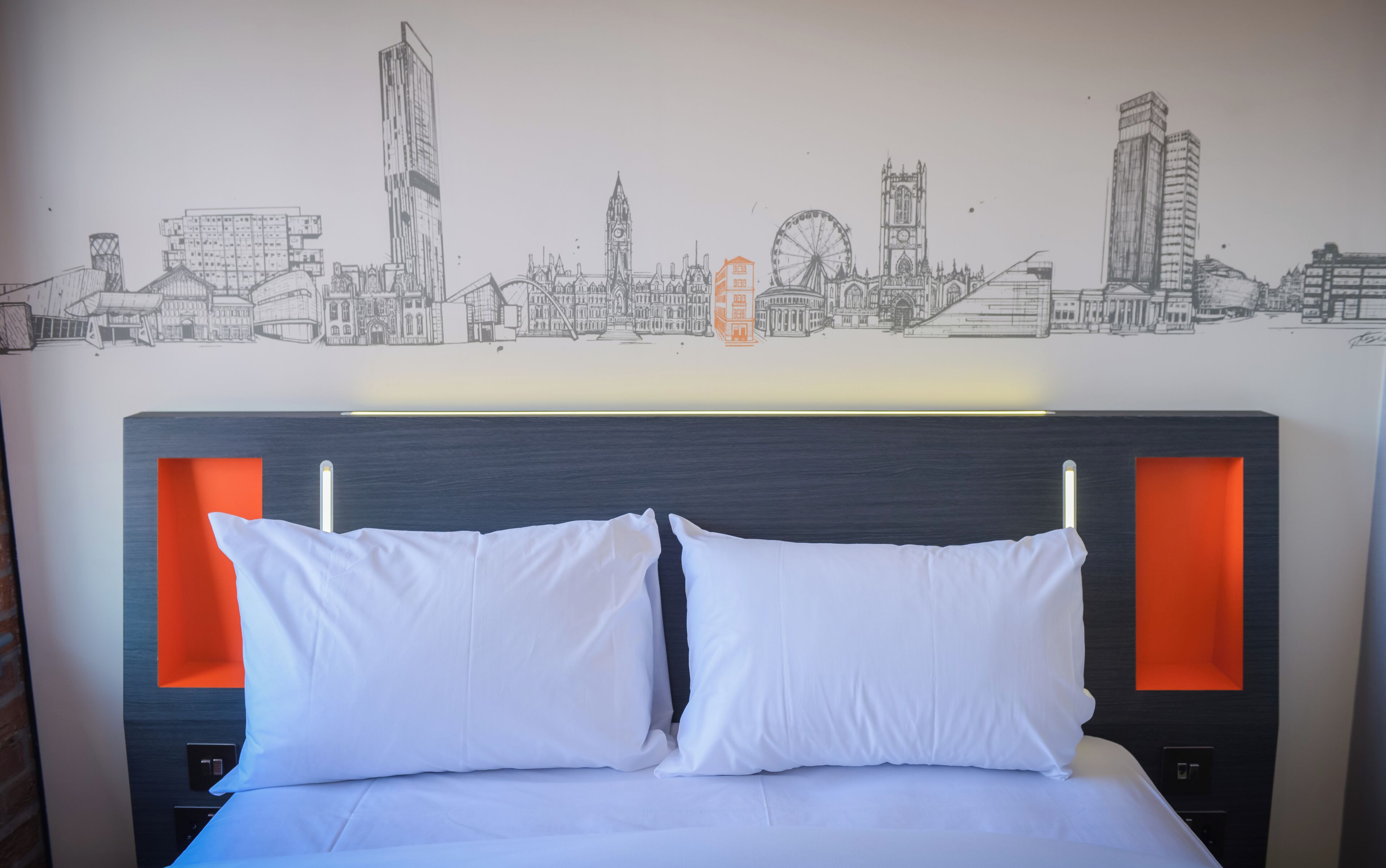 Ein Hotelzimmer mit Bett, weißen Kissen und Wandgemälde mit Stadtansicht.