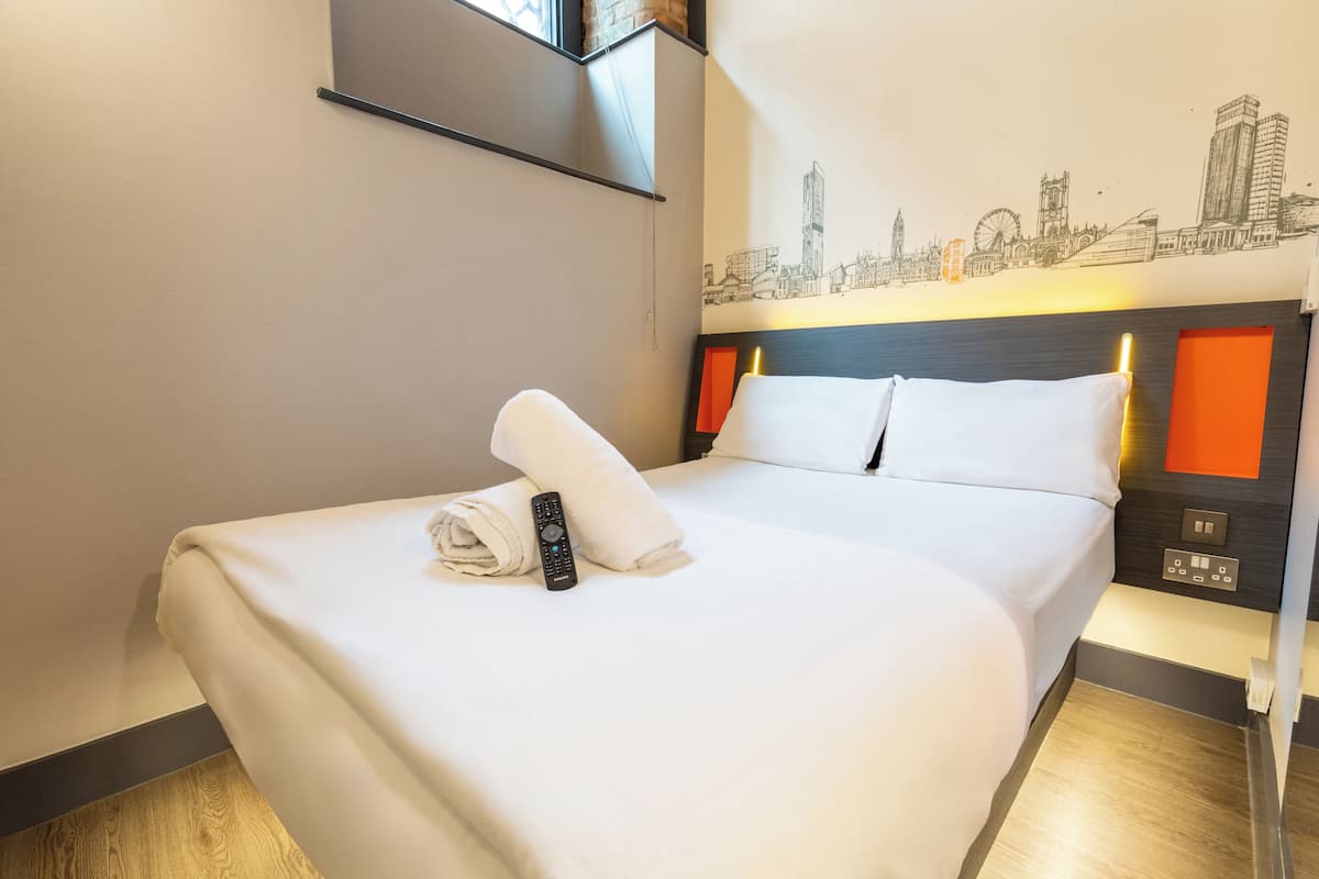 A hotel room with a neatly made bed, a remote control, and a cityscape mural on the wall.