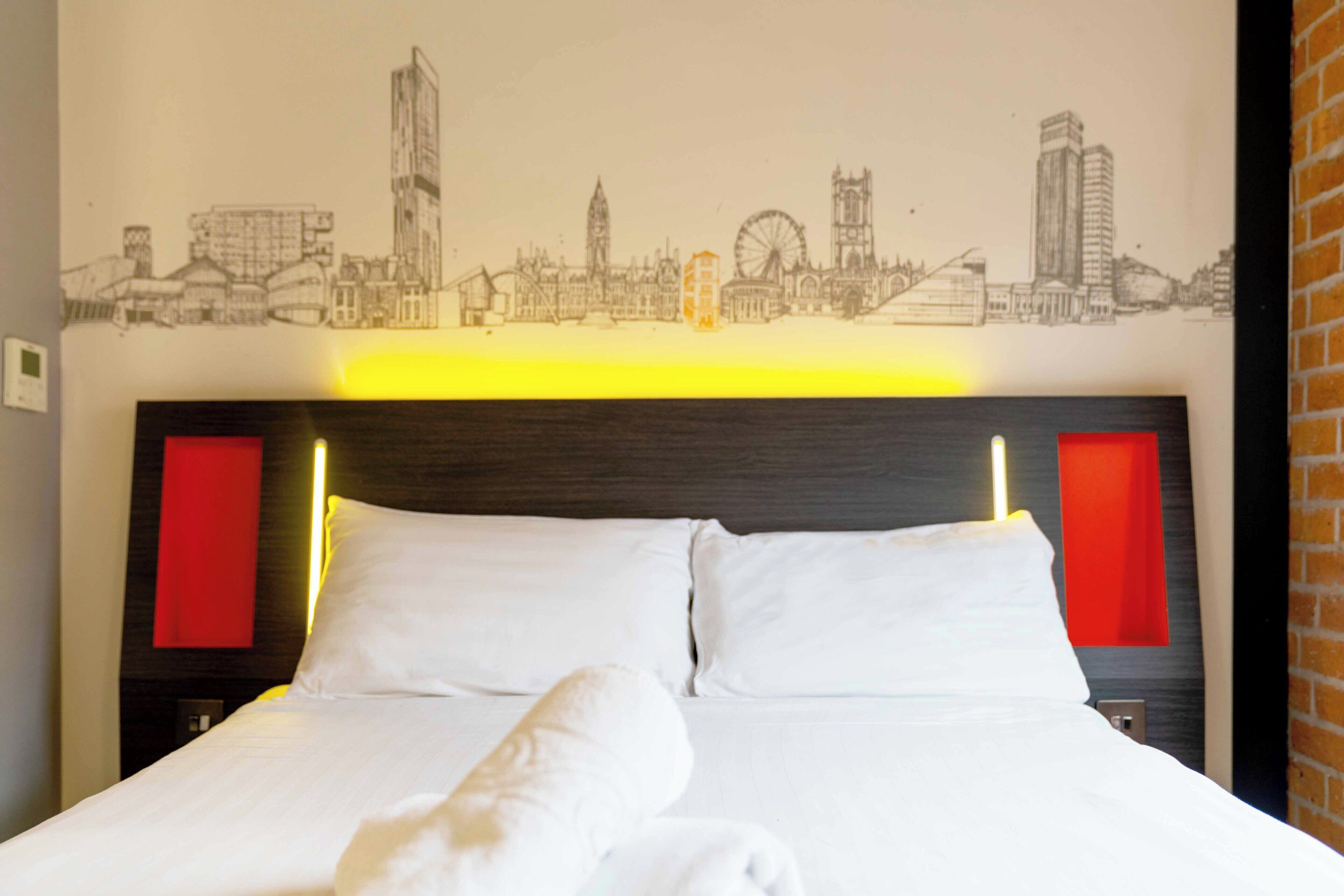 A hotel room with a bed, a cityscape mural on the wall, and red accent lighting.