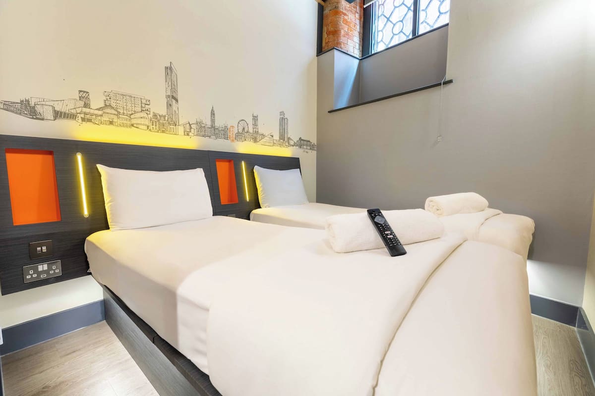 A hotel room with two beds, a cityscape wall art, a TV mounted on the wall, and a window with a view.