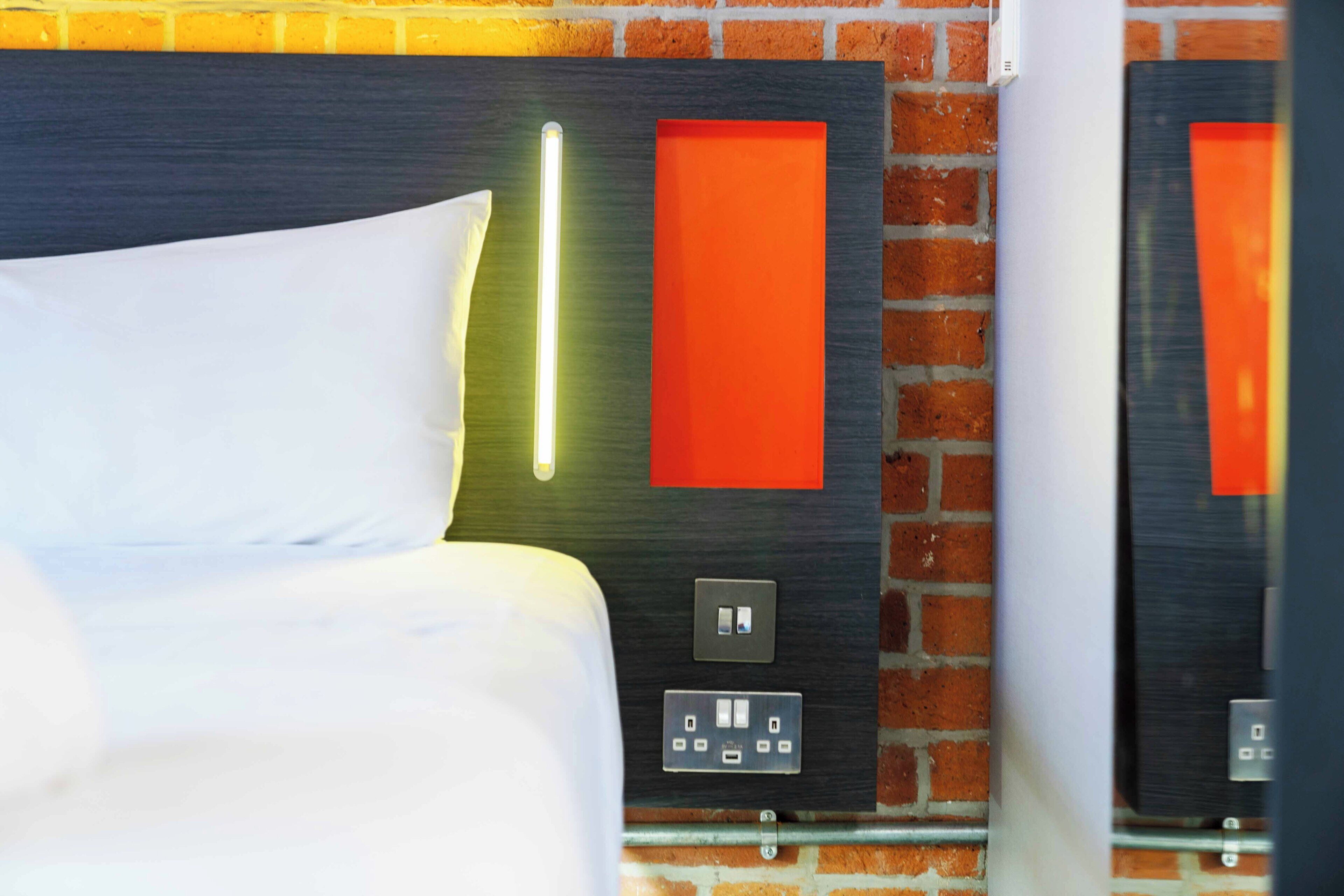 A hotel room with a bed, a wall-mounted light, a red accent wall, and a power outlet.