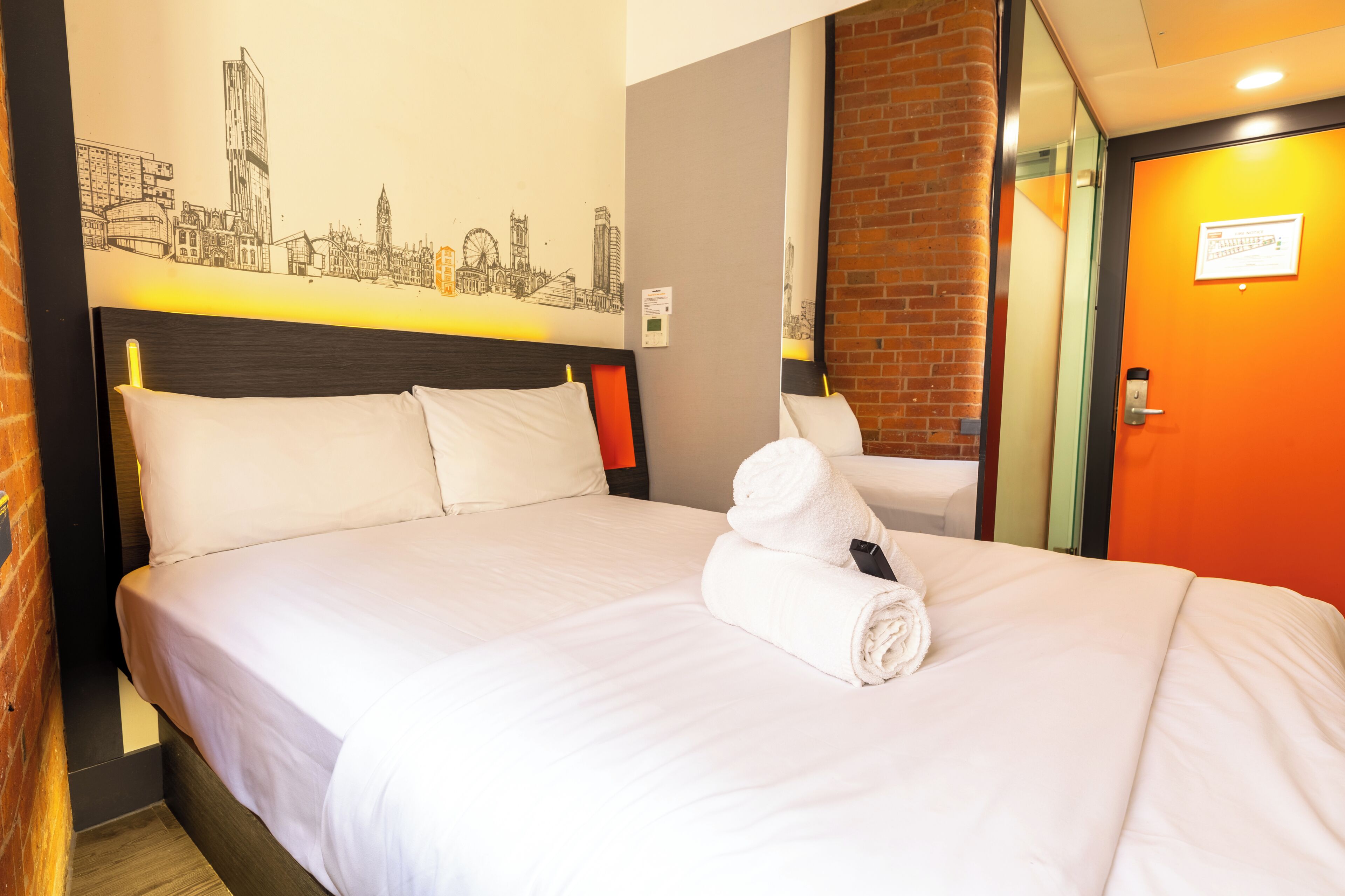Ein Hotelzimmer mit Bett, Kissen, einem zusammengerollten Handtuch, einer Stadtlandschaft als Wandbild und einer Ziegelwand.