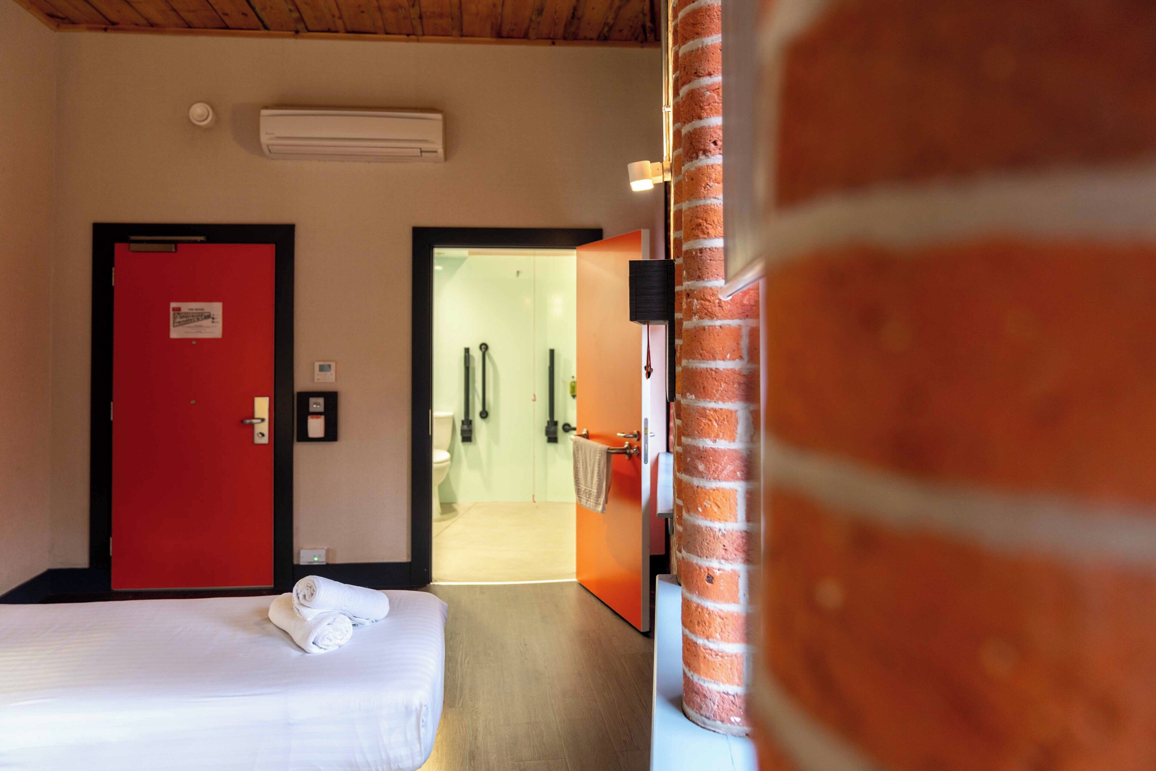 Une chambre d’hôtel avec une porte rouge, un lit avec du linge de lit blanc, une colonne en briques et une salle de bain visible.