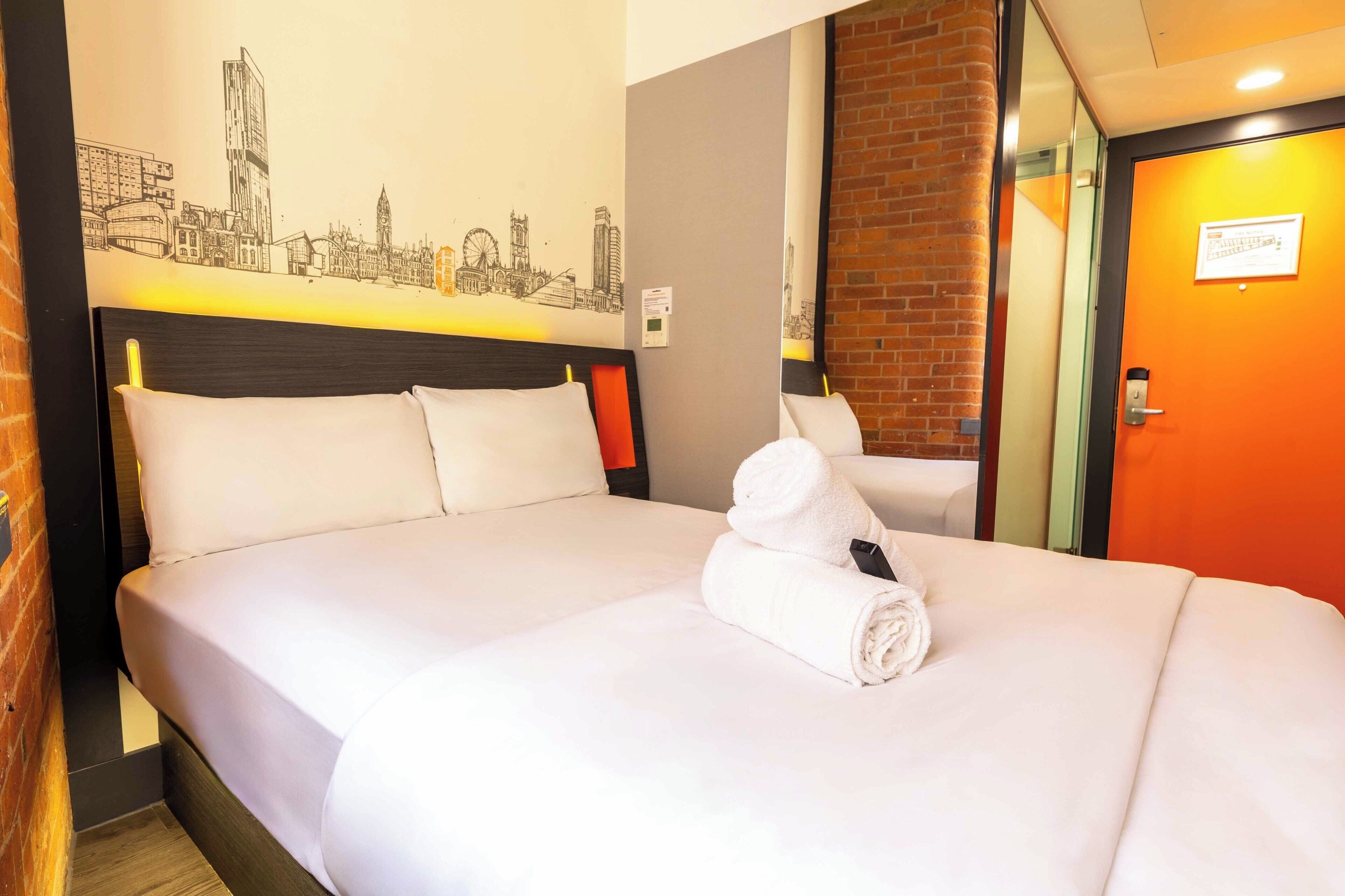 A hotel room with a bed, pillows, a rolled towel, a cityscape mural, and a brick accent wall.