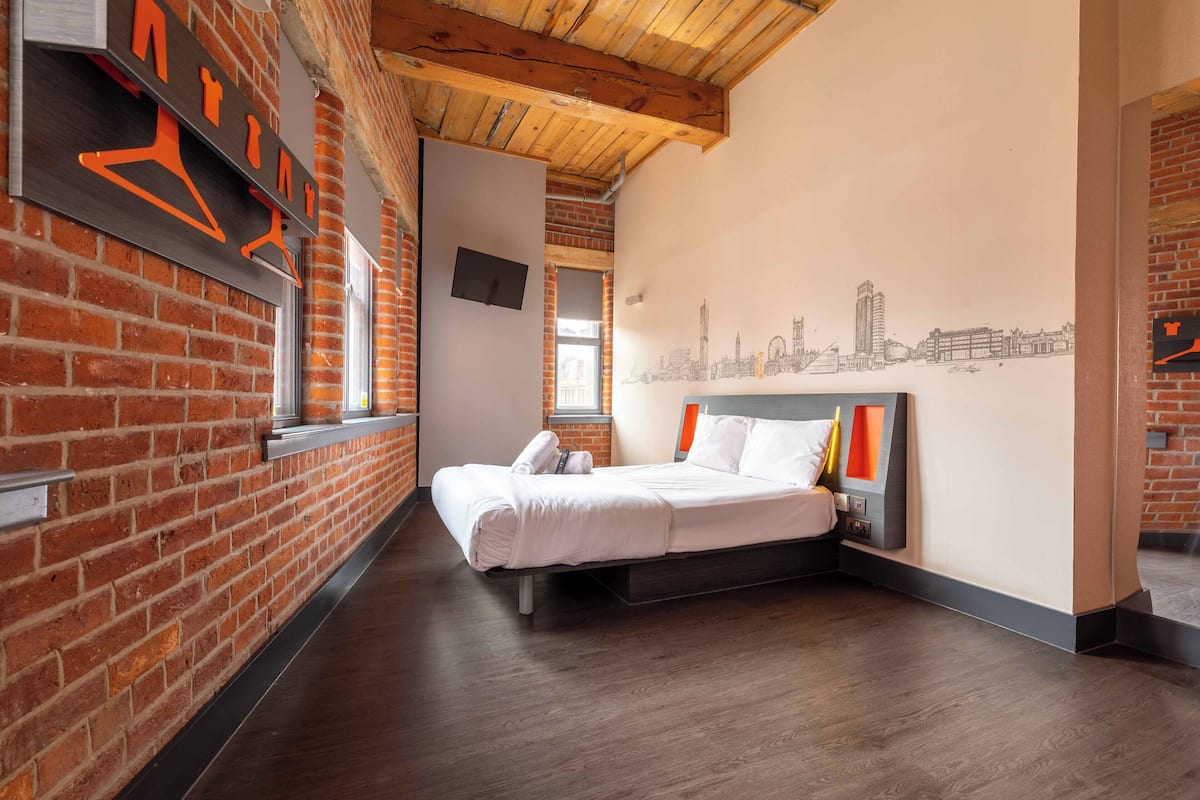 A hotel room with a brick wall, a bed with white bedding, a cityscape mural, and a wooden ceiling.