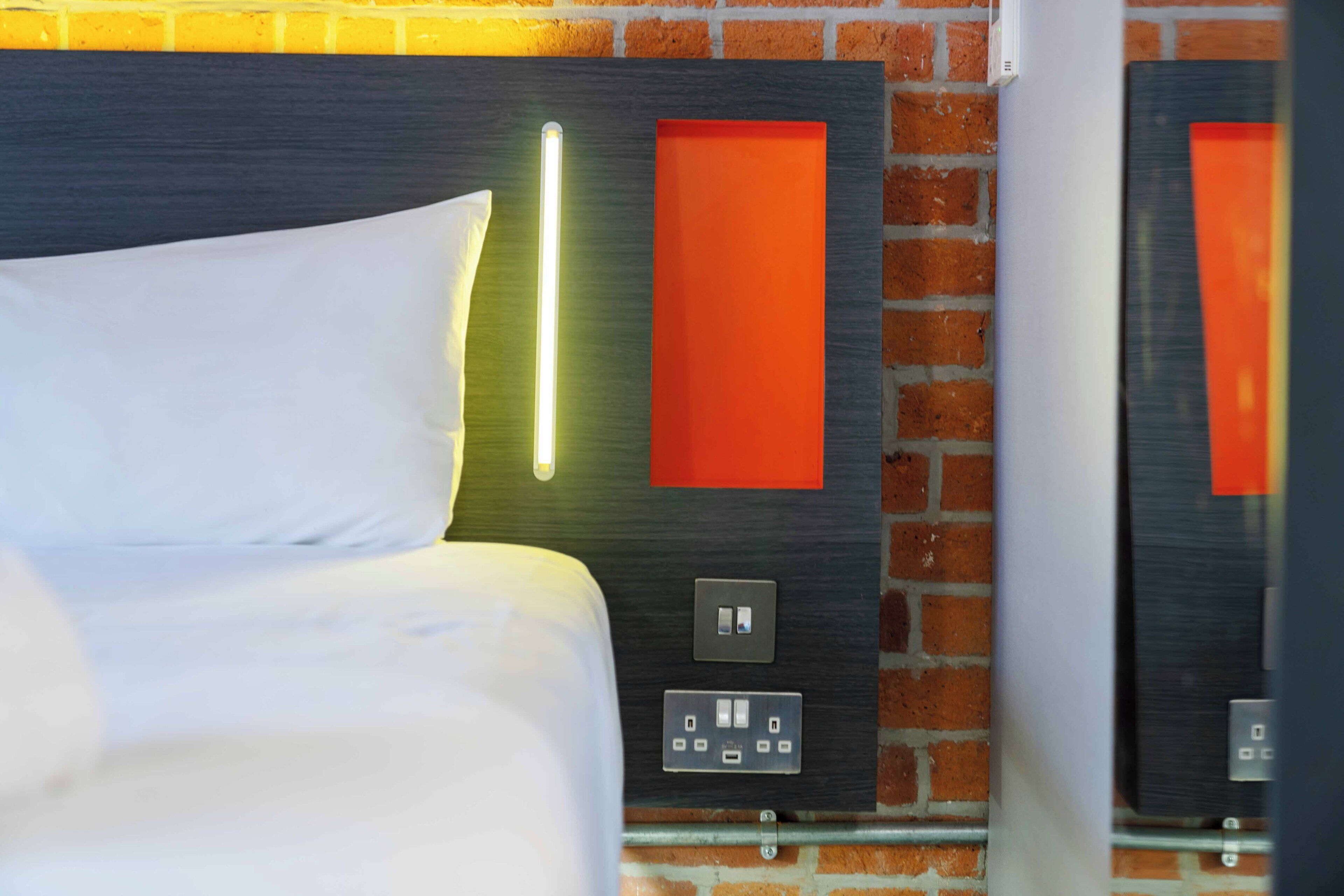 A hotel room with a bed, a wall-mounted light fixture, a power outlet, and a red accent wall.