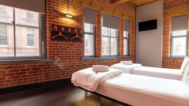 A room with two beds, a brick wall, large windows, and a hanging garment rack. - easyHotel Manchester (Manchester)