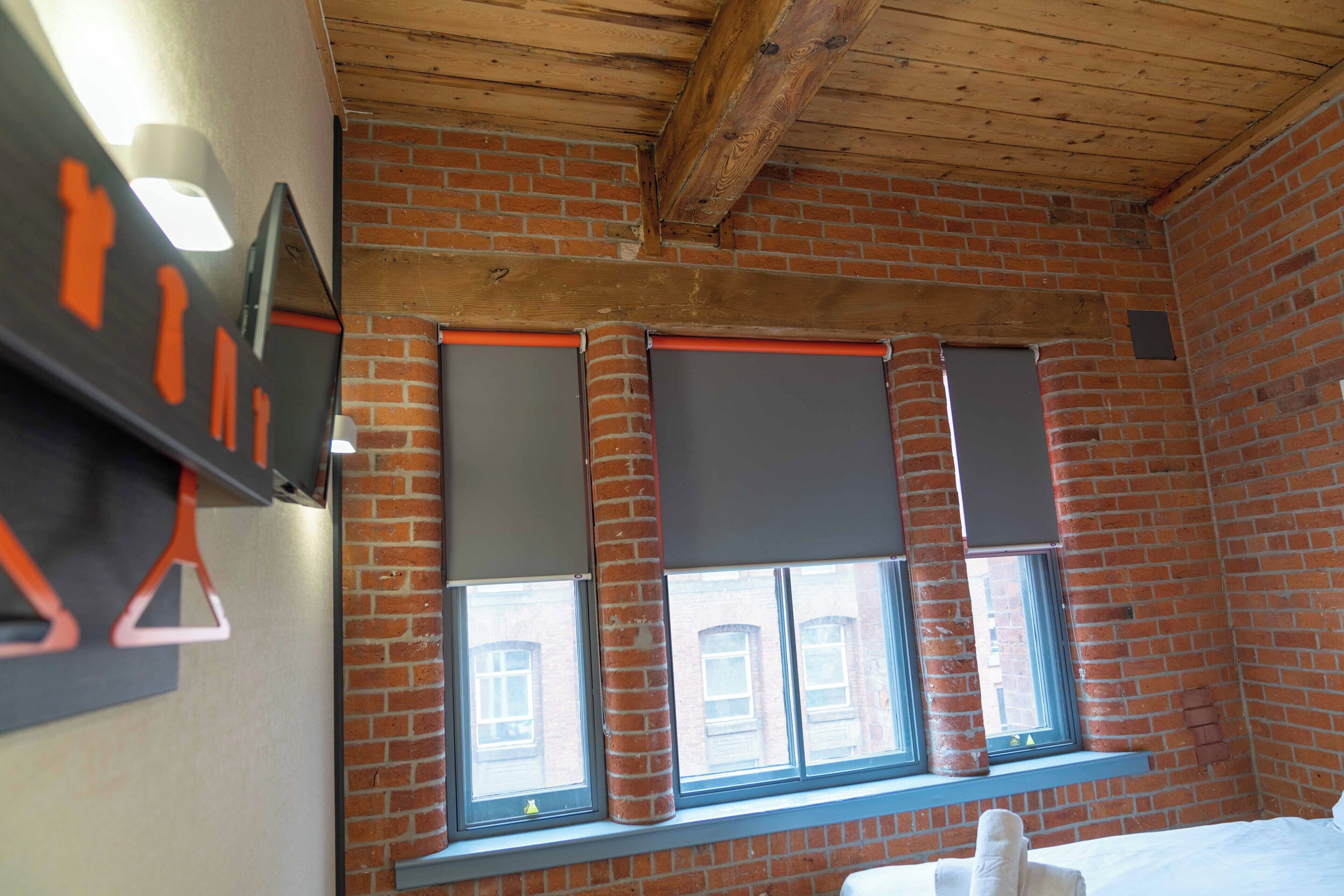 A room with brick walls, wooden beams, and large windows with blinds.