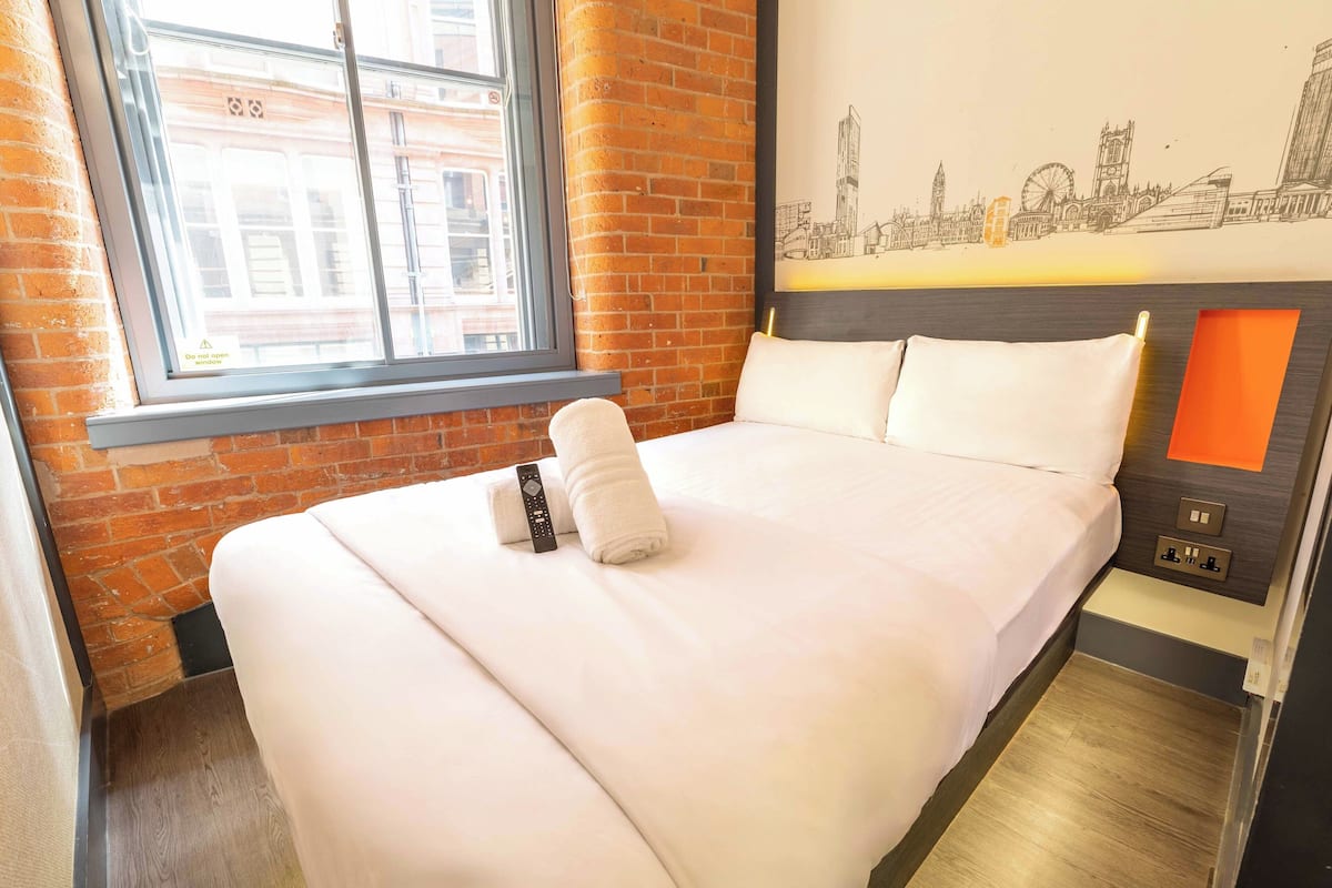A hotel room with a brick wall, a large window, a bed with white linens, a remote control, and a wall-mounted control panel.