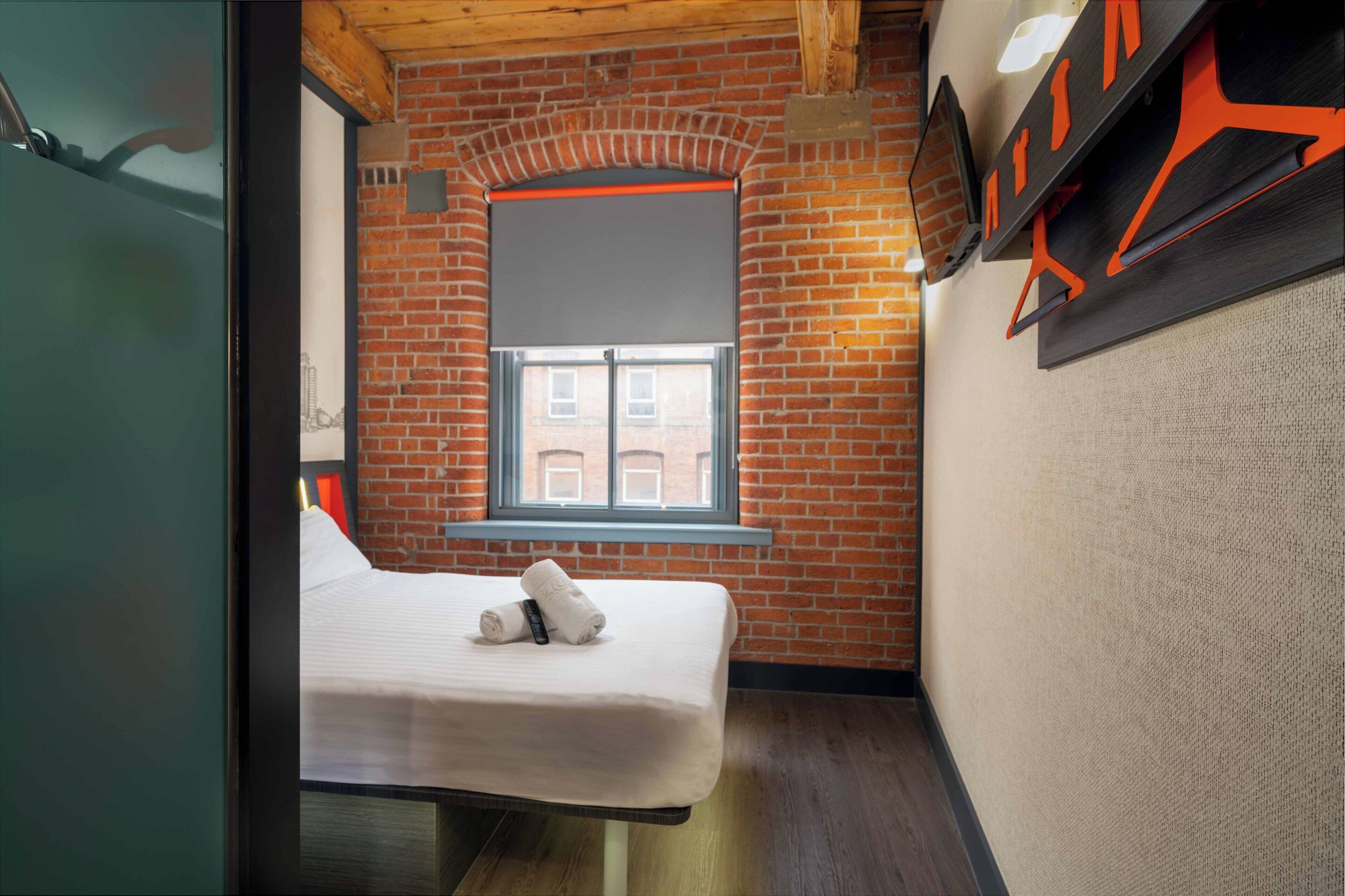 A compact room with a brick wall, a window, a bed, and a wall-mounted TV.