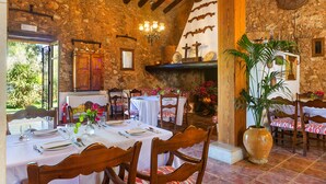 Un comedor rústico con paredes de piedra, vigas de madera y una araña. Las mesas están dispuestas con manteles blancos y sillas.