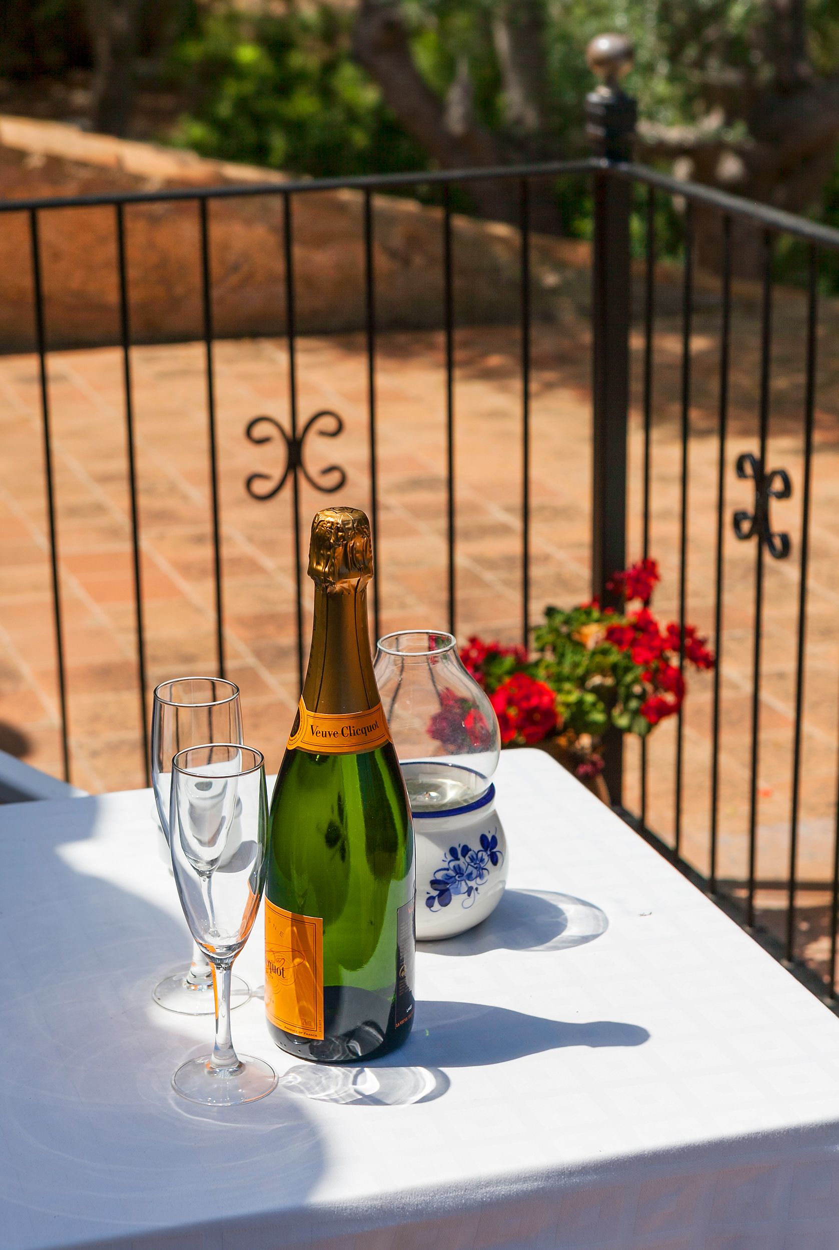 Una mesa con una botella de champán, dos copas y un jarrón decorativo en una terraza con barandilla y flores al fondo.
