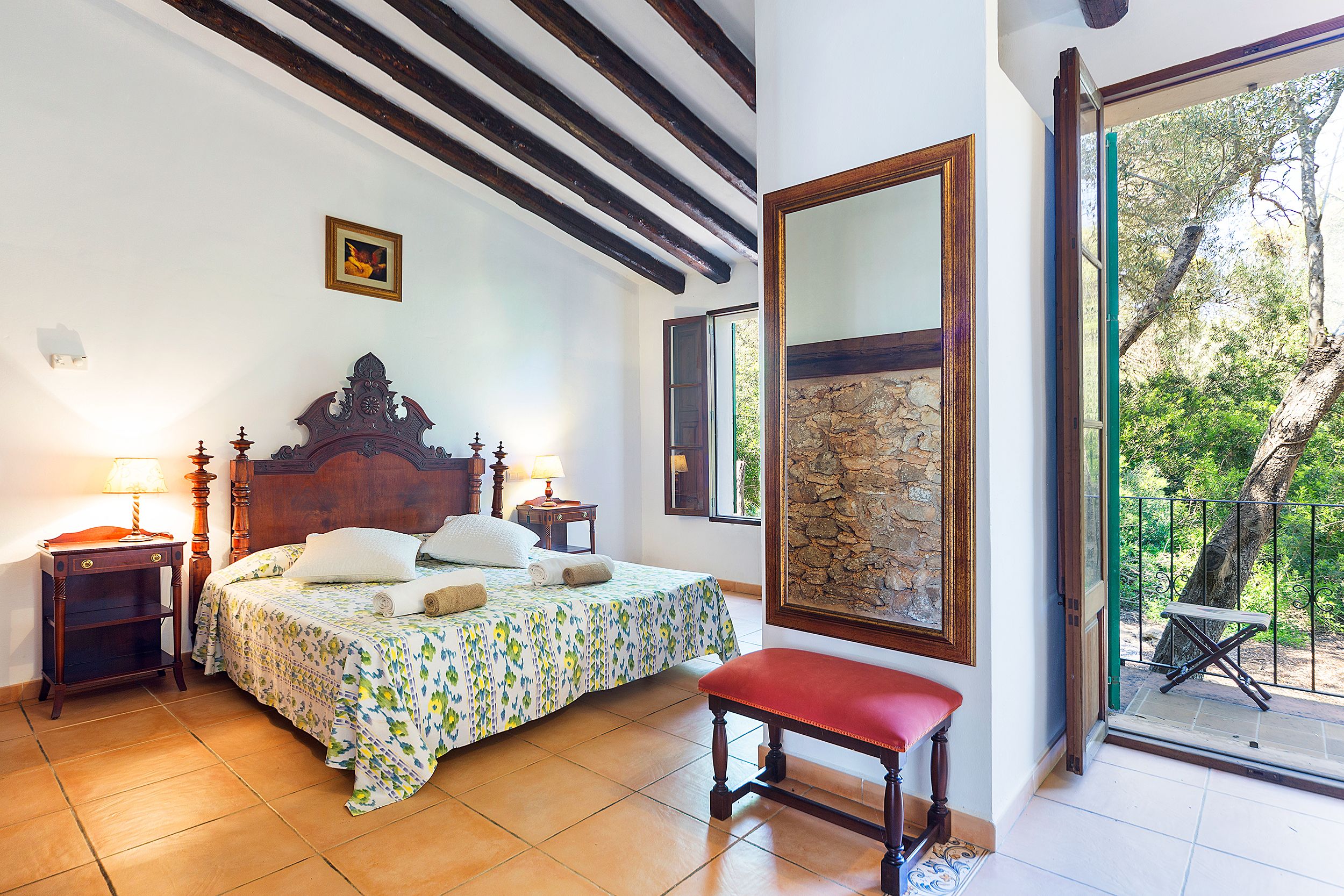 A bedroom with a wooden bed, a bench, a nightstand, and a large window with a view of trees.