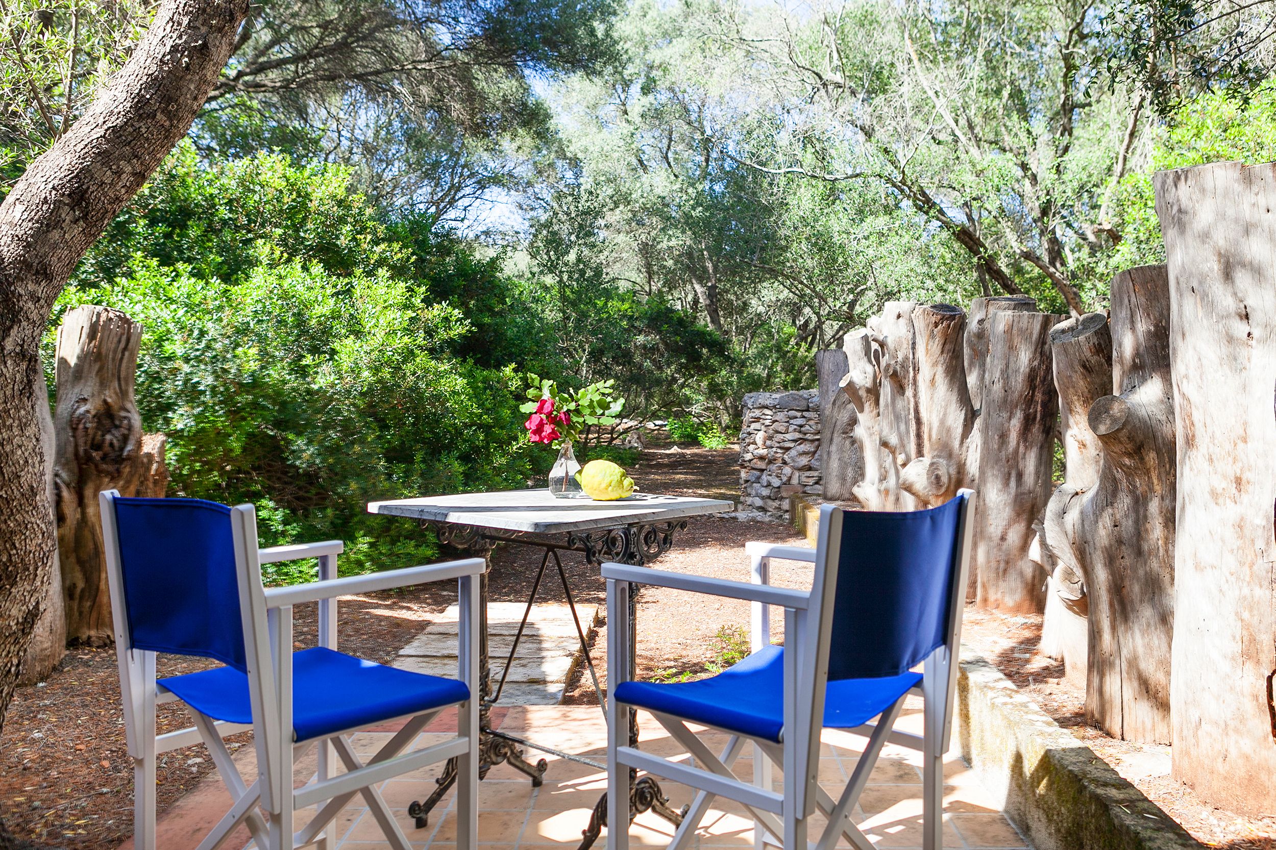 An outdoor seating area with two blue chairs and a table, surrounded by trees and stone walls.