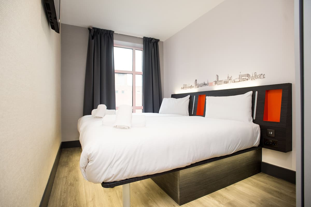 A modern hotel room with a large bed, a cityscape wall art, and a window with curtains.