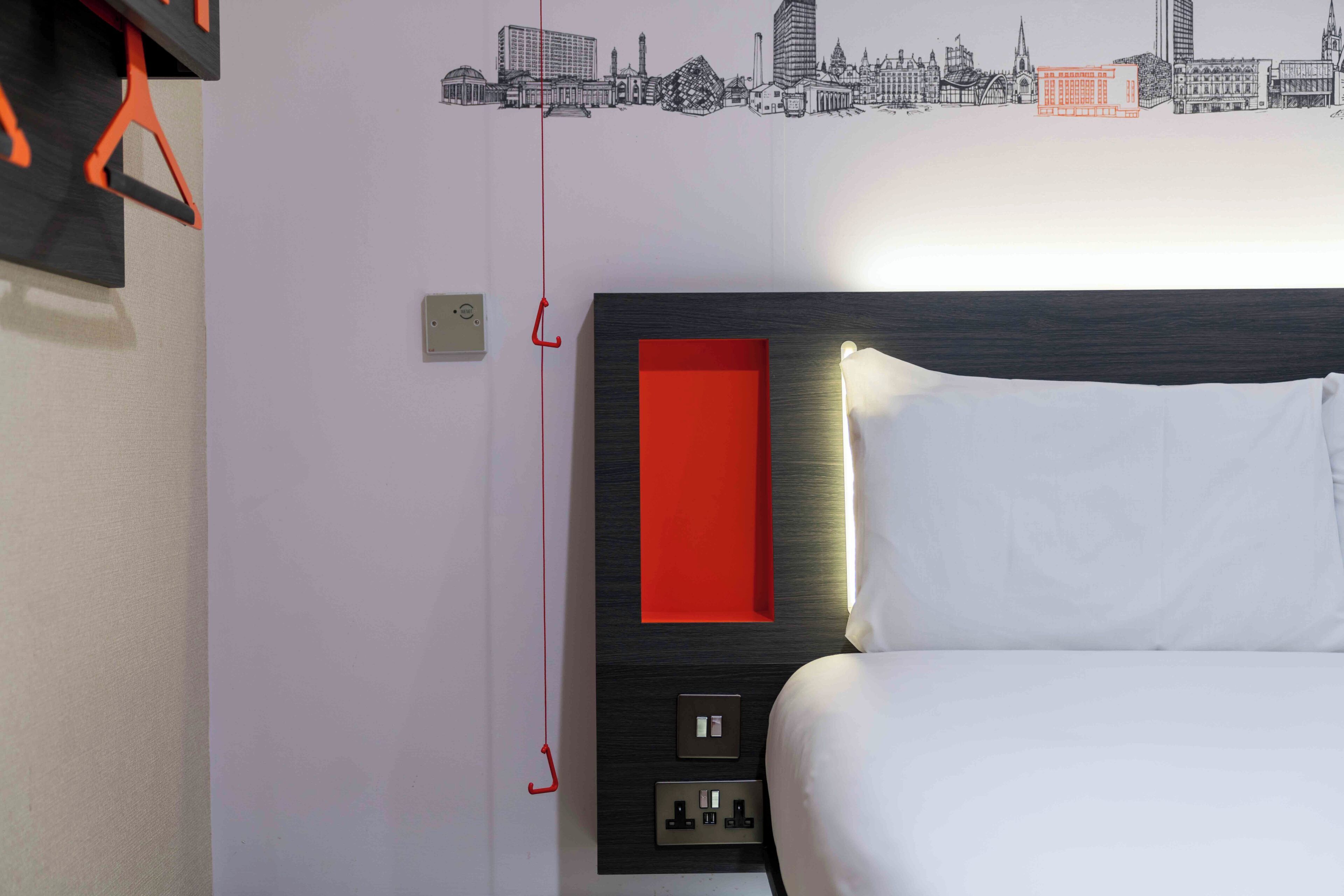 A hotel room with a bed, a cityscape wall art, a red accent wall, and a power outlet.