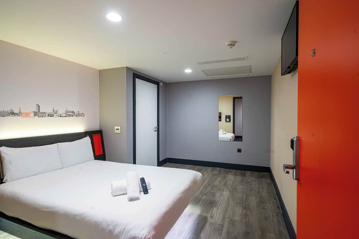 A hotel room with a bed, a wall-mounted TV, a mirror, and a cityscape artwork.