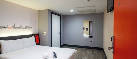 A hotel room with a bed, a wall-mounted TV, a mirror, and a cityscape artwork.