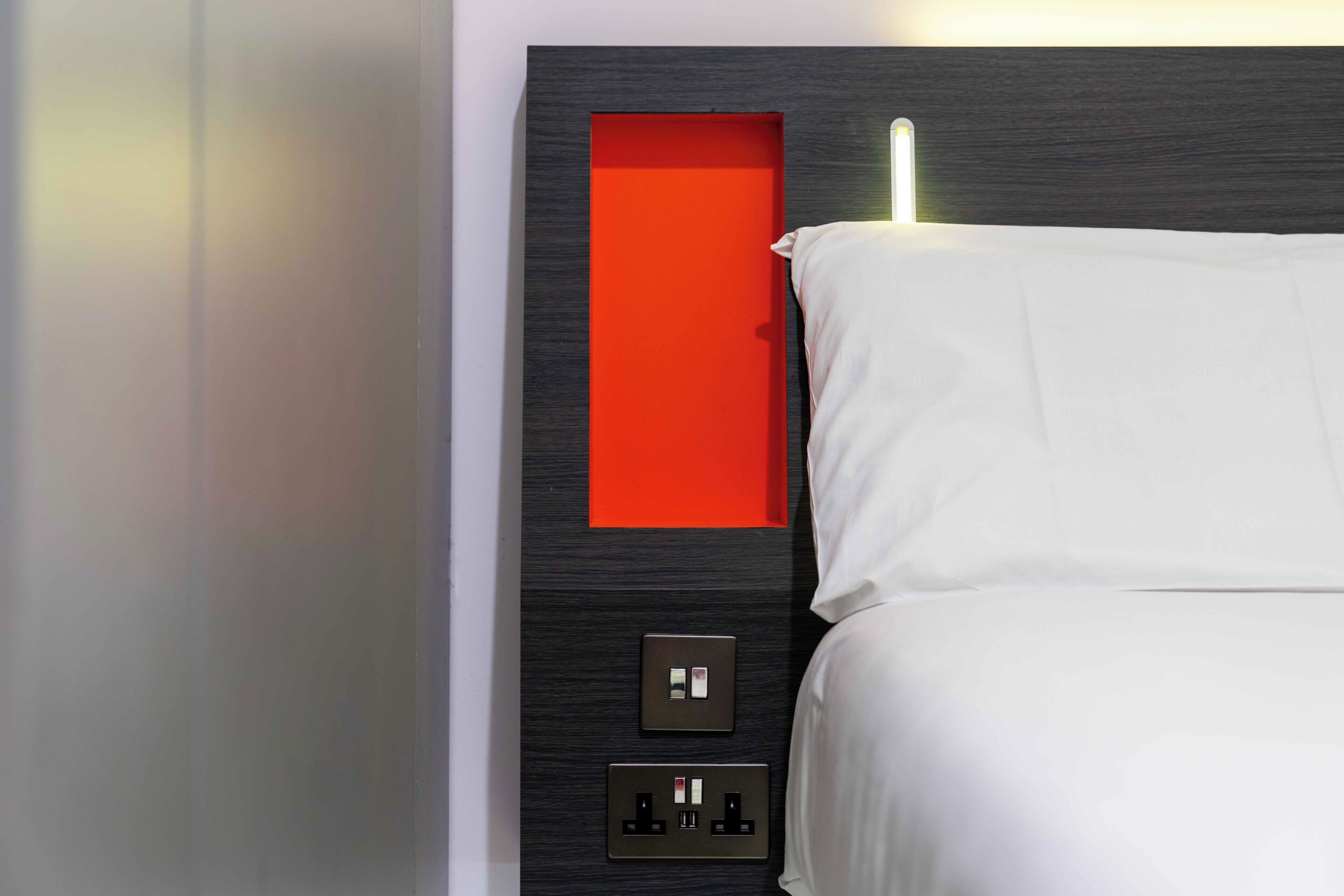 Une chambre d’hôtel moderne avec une tête de lit en bois foncé, un panneau décoratif rouge, un lit blanc et un panneau de commande mural doté de deux interrupteurs et d’une prise.