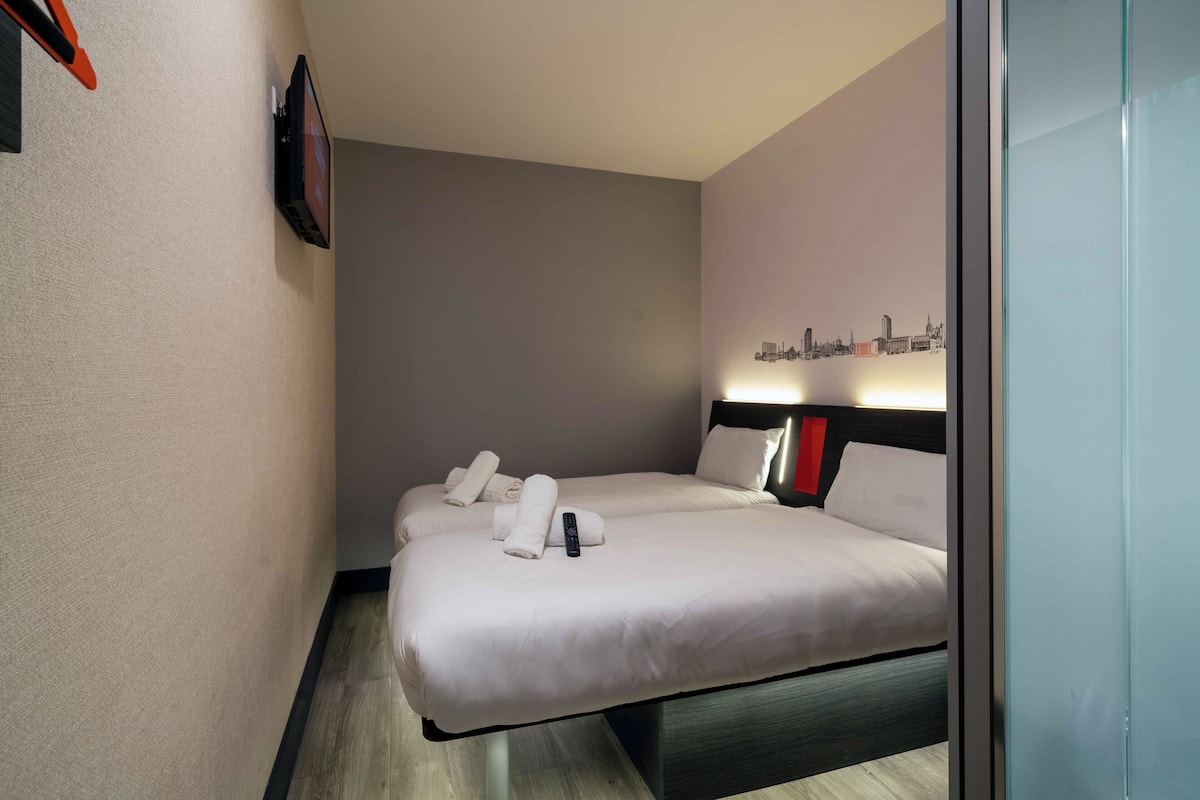 A hotel room with two beds, a flat-screen TV mounted on the wall, and a cityscape artwork on the wall.