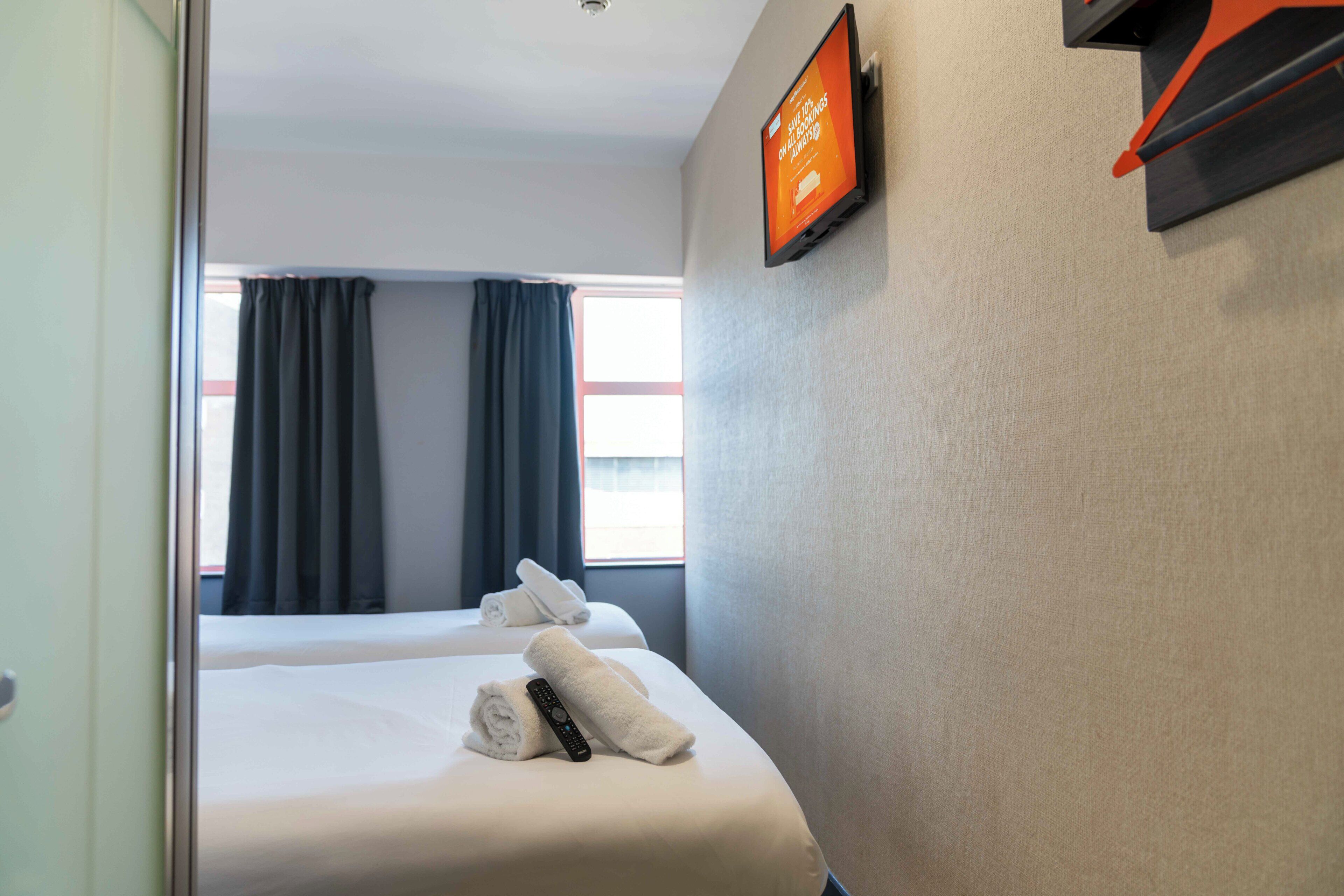 A hotel room with two beds, a TV mounted on the wall, and a window with curtains.