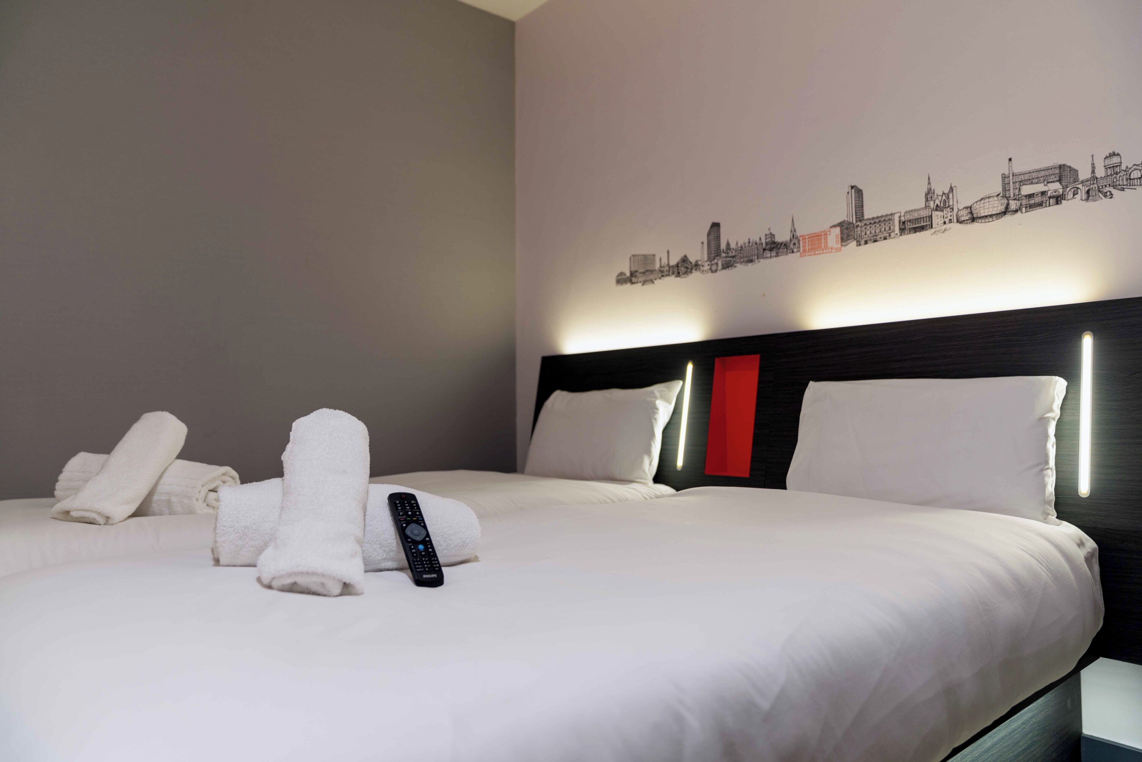 Une chambre d’hôtel avec un lit parfaitement fait, une télécommande et une décoration murale représentant un paysage urbain.