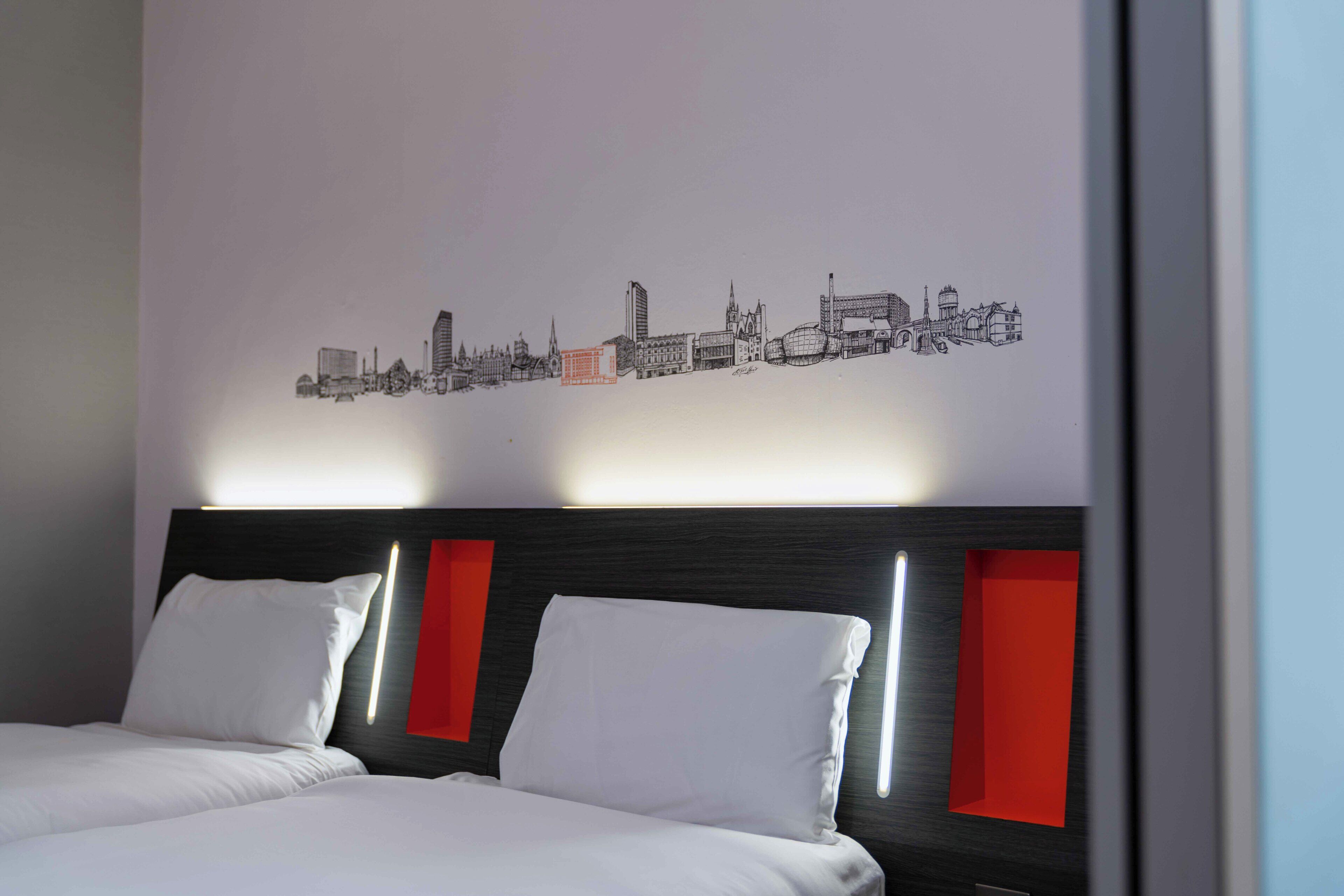 A hotel room with a bed, two pillows, a headboard with red accents, and a cityscape wall art.