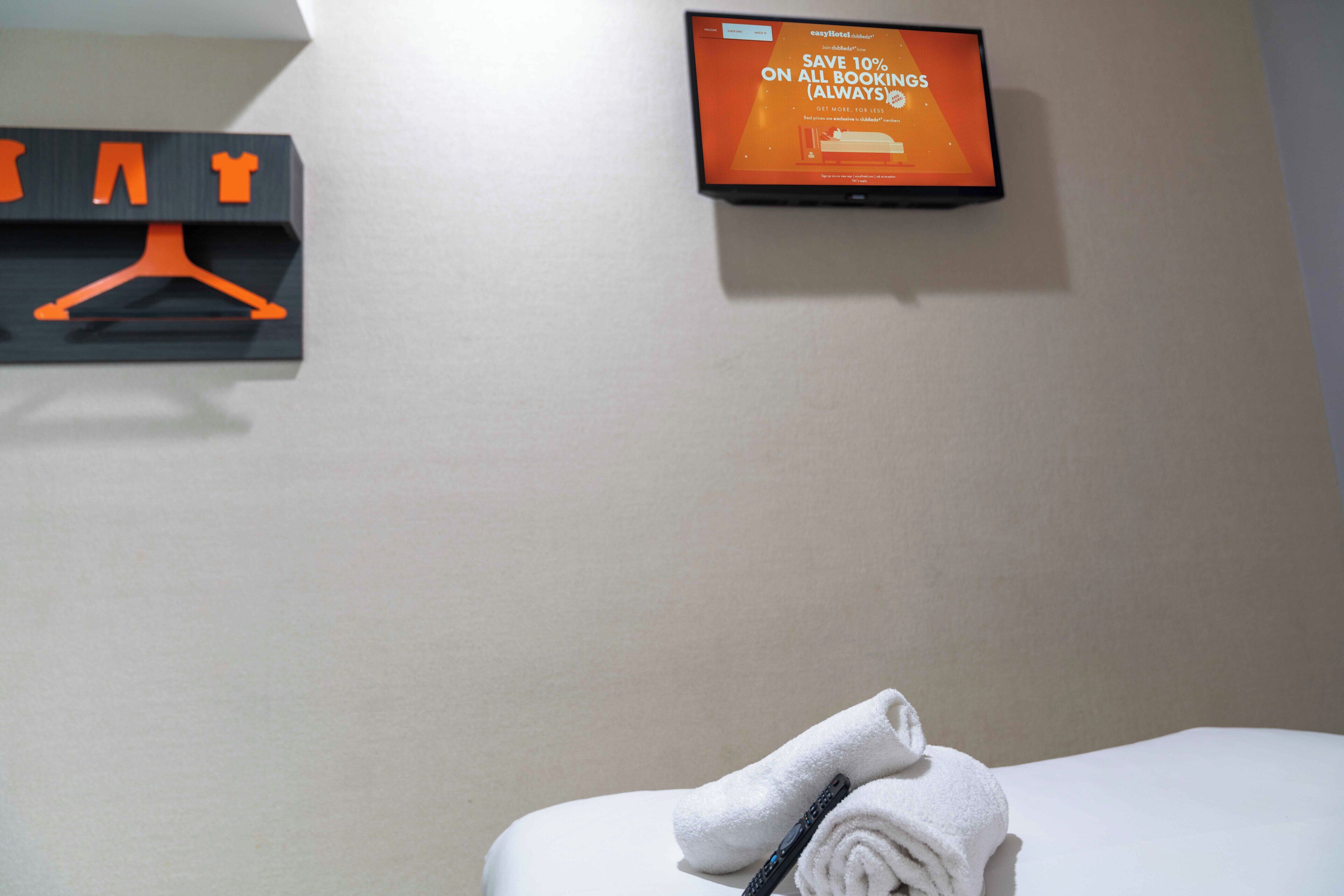 Quarto de hotel com cama, televisão na parede exibindo uma mensagem promocional e um cabideiro com cabides.
