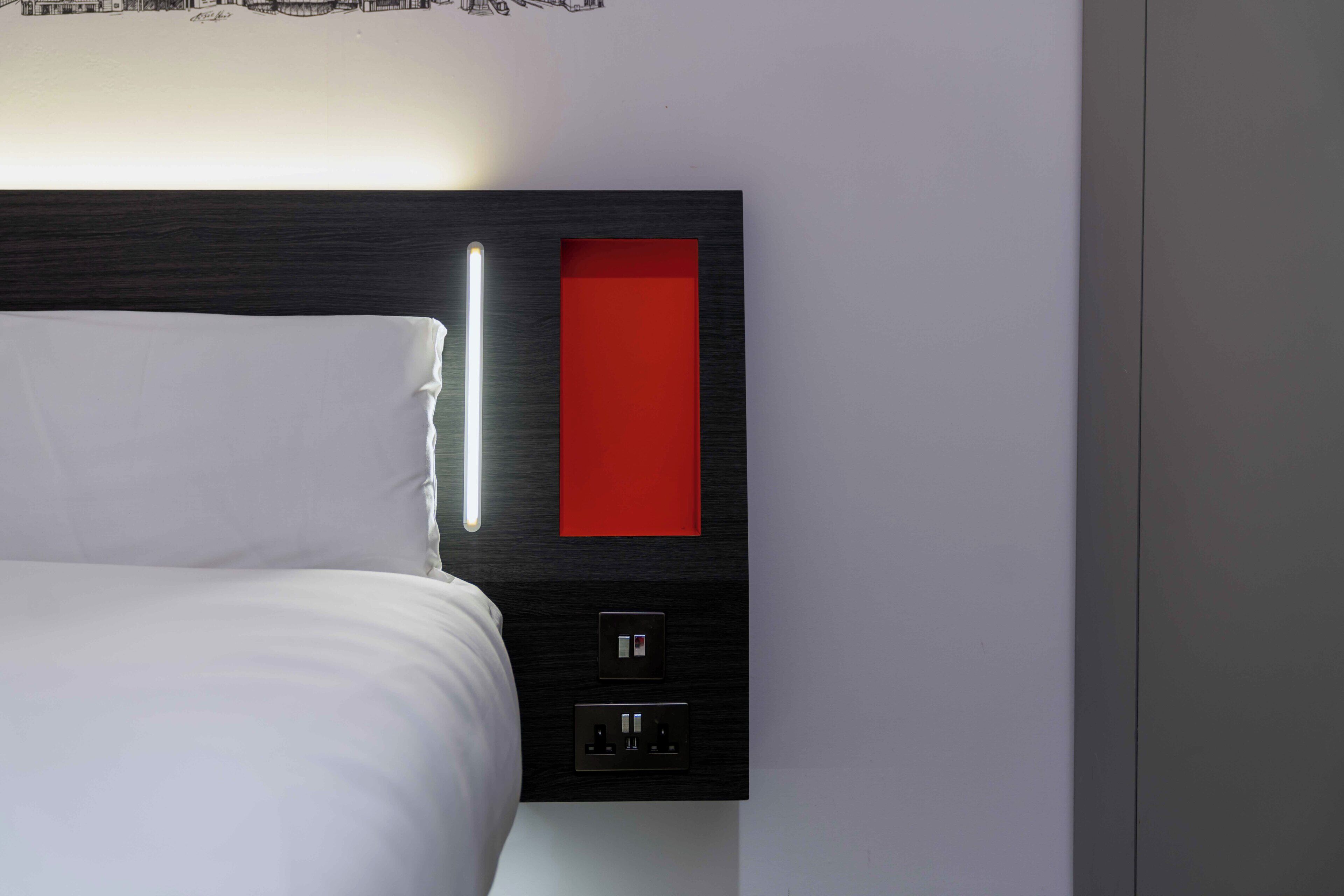 A modern hotel room with a bed, a red accent panel, and a wall-mounted control panel.
