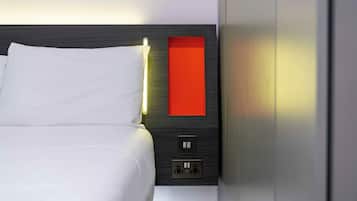 A neatly made bed with white linens, a red accent panel on the headboard, and a wall-mounted light fixture.